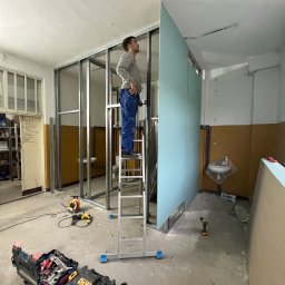 Aliaksandr Filipau - Pracownik na drabinie montuje płytę gipsowo-kartonową do metalowego stelaża w pomieszczeniu w trakcie remontu, widoczne narzędzia i skrzynki narzędziowe.