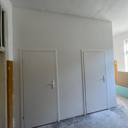 Aliaksandr Filipau - Wnętrze po odświeżeniu: Dwie białe, proste drzwi na tle świeżo pomalowanej ściany w jasnym odcieniu. Widoczne nierówności na suficie. Podłoga betonowa, częściowo pokryta resztkami po remoncie.