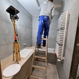 Aliaksandr Filipau - Pracownik na drewnianej drabinie, w niebieskich spodniach roboczych i szarej bluzie z kapturem, poprawia gładź na suficie w łazience z szarymi płytkami, widoczny grzejnik drabinkowy i lampa...