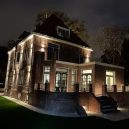 Oświetlona nocą, dwukondygnacyjna, ceglana rezydencja z czerwonym dachem i czarnymi ramami okiennymi, otoczona zielonym trawnikiem.