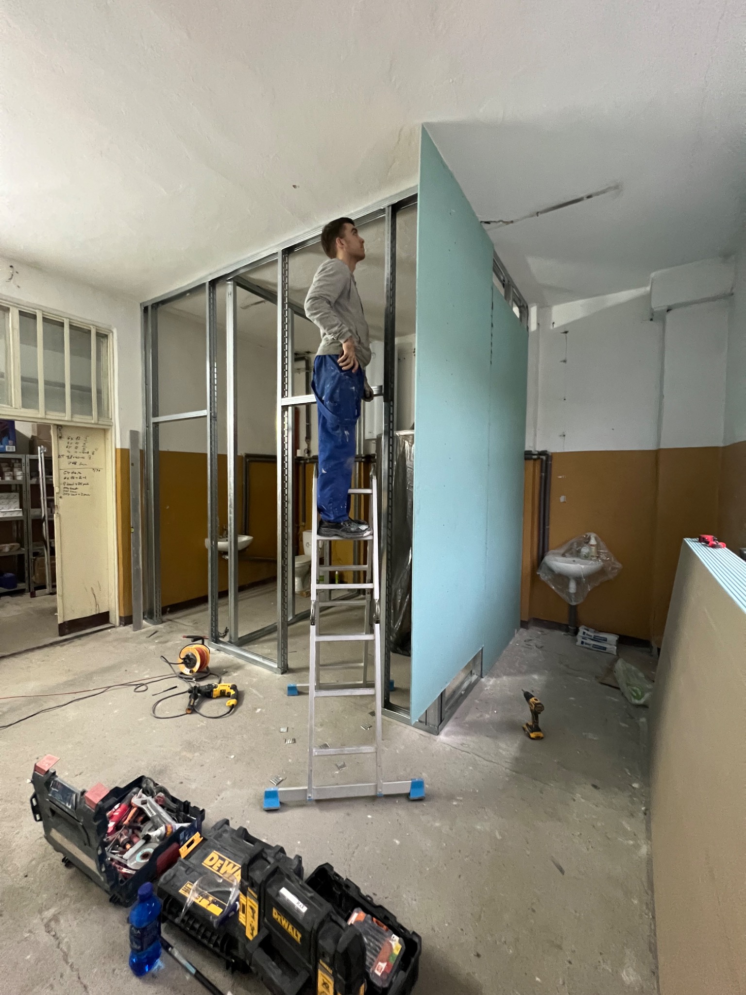 Pracownik na drabinie montuje płytę gipsowo-kartonową do metalowego stelaża w pomieszczeniu w trakcie remontu, widoczne narzędzia i skrzynki narzędziowe.