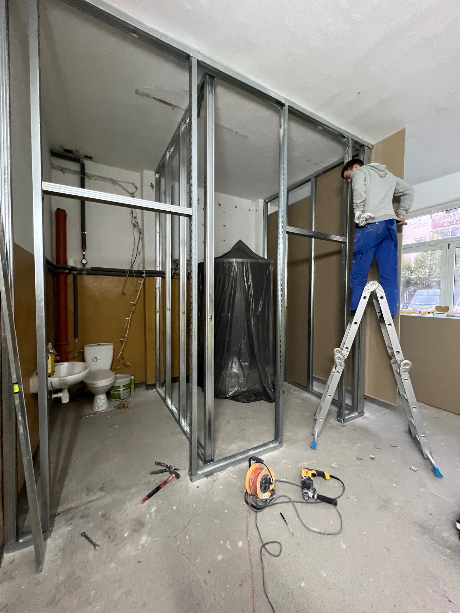 Montaż ścianki działowej z płyt gipsowo-kartonowych na metalowym stelażu w łazience, widoczny pracownik na drabinie, narzędzia na podłodze i sanitariaty w tle.