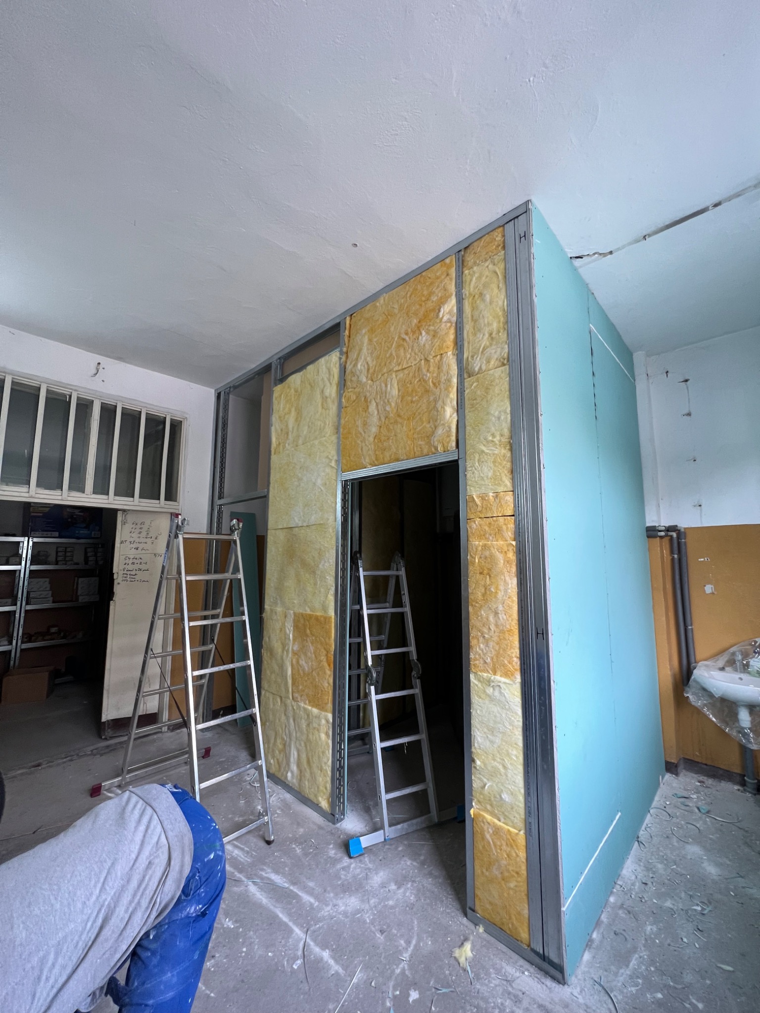 Stelaż metalowy z izolacją z wełny mineralnej i płytą gipsowo-kartonową w trakcie montażu, widoczne drabiny i fragment sylwetki pracownika w kombinezonie.