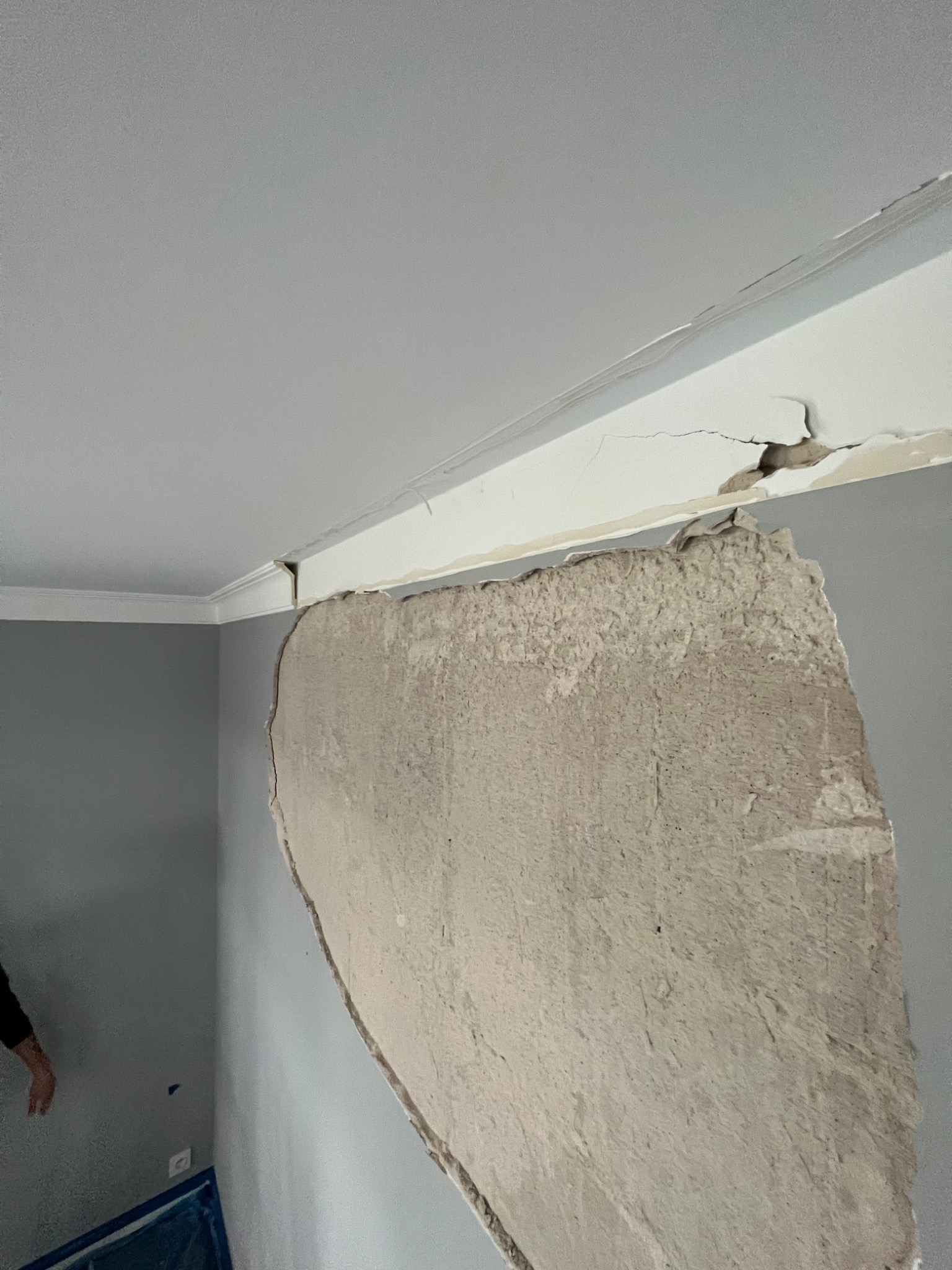 Uszkodzona ściana w rogu pokoju z odpadającym tynkiem, widoczne pęknięcia na listwie sufitowej i szary kolor ścian.