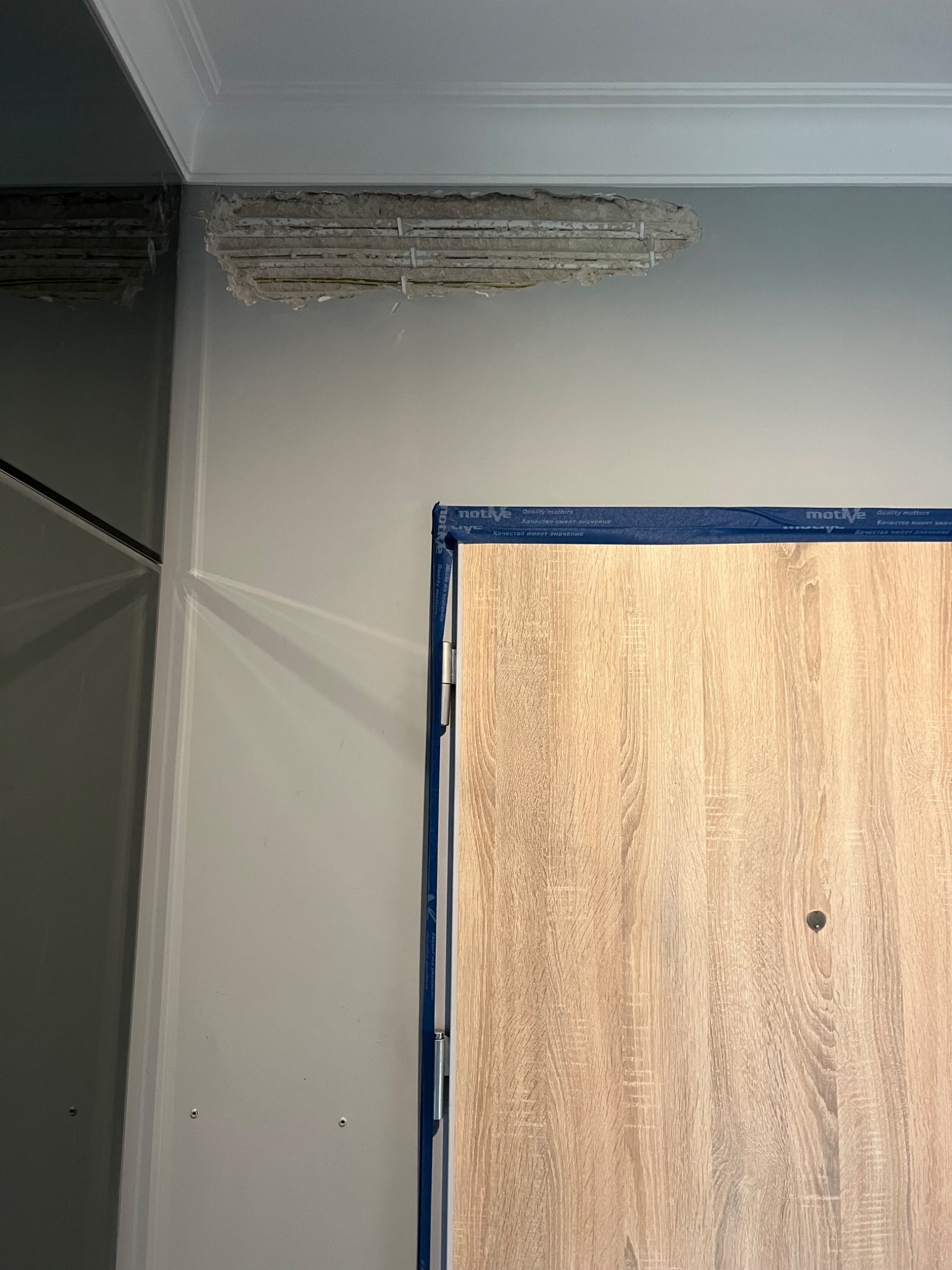 Fragment ściany z uszkodzonym narożnikiem sufitu, odsłaniającym konstrukcję, oraz fragment zabezpieczonych taśmą drzwi o wyglądzie drewna.