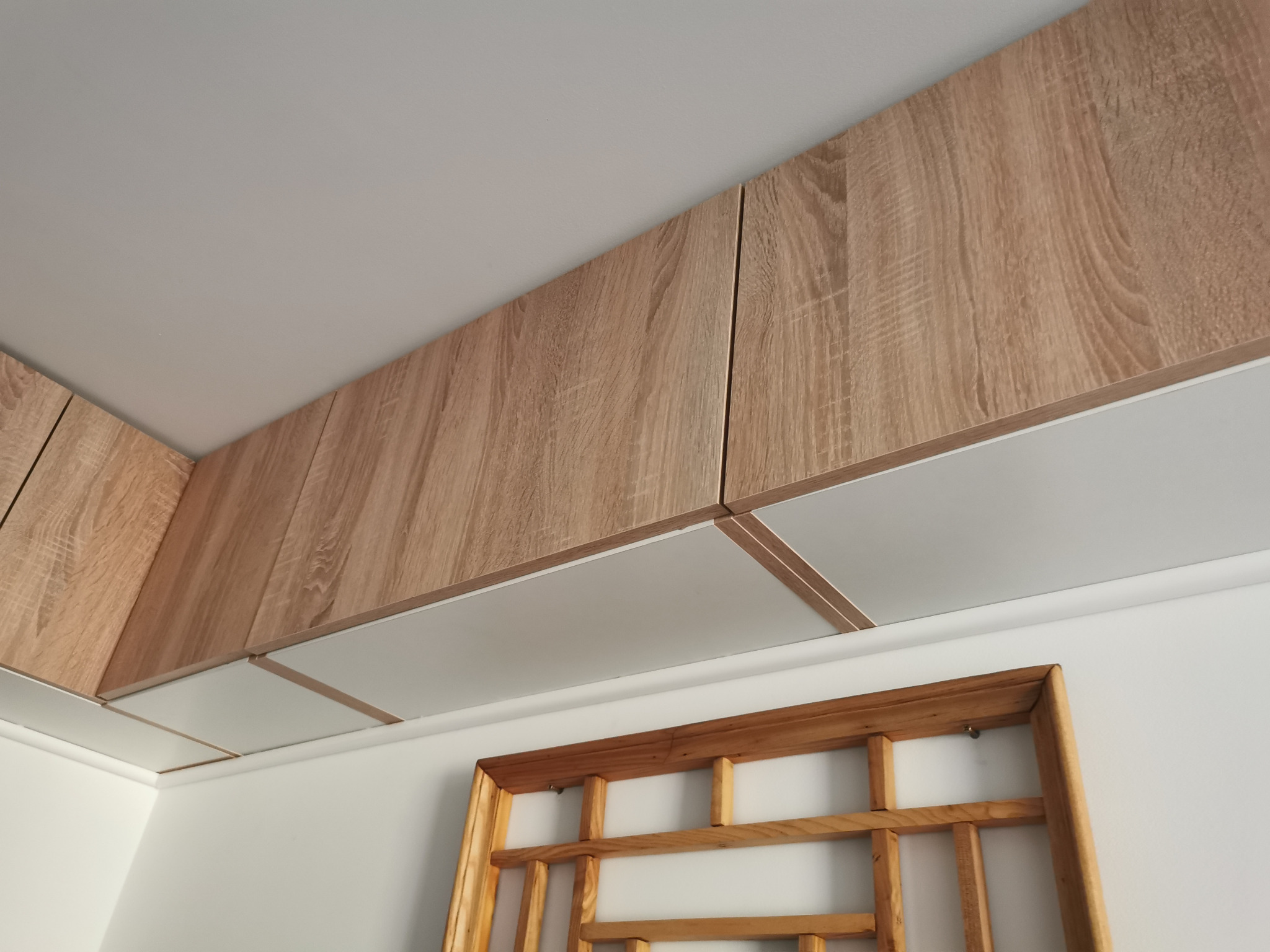 Zamontowane pod sufitem szafki kuchenne z drewnopodobnymi frontami i białymi spodami, widoczne fragmenty ścian i dekoracyjna drewniana rama na ścianie.