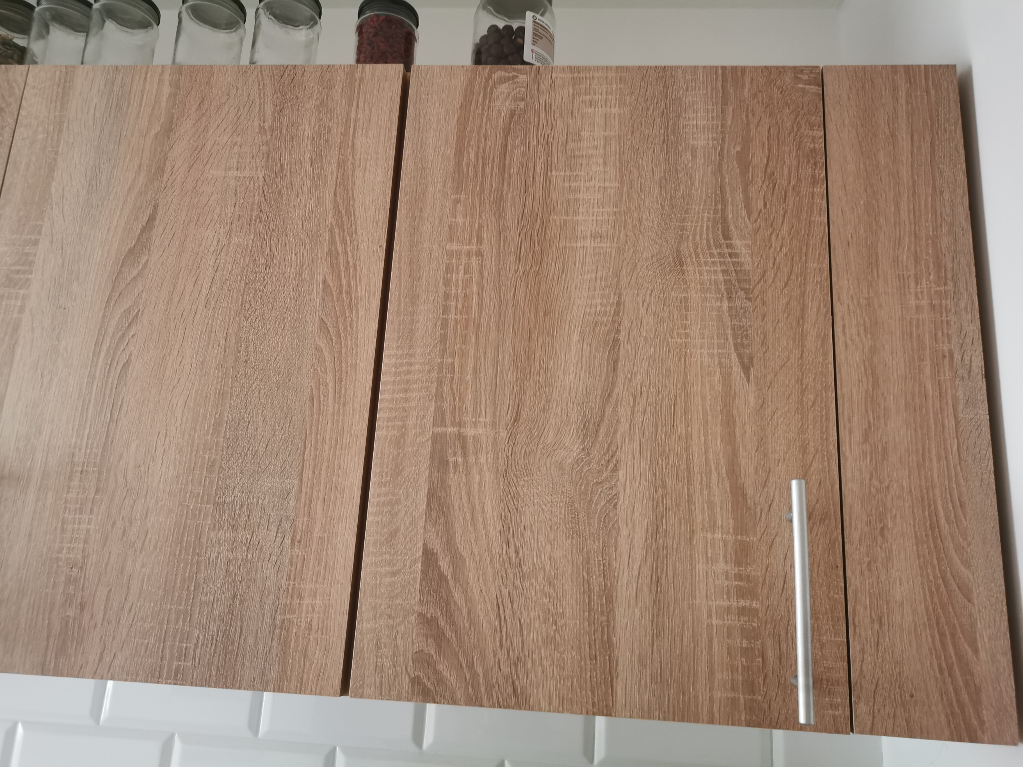 Zamknięte szafki kuchenne z drewnopodobnym wykończeniem, srebrne uchwyty, na górze widoczne szklane pojemniki z przyprawami, białe płytki na ścianie poniżej.