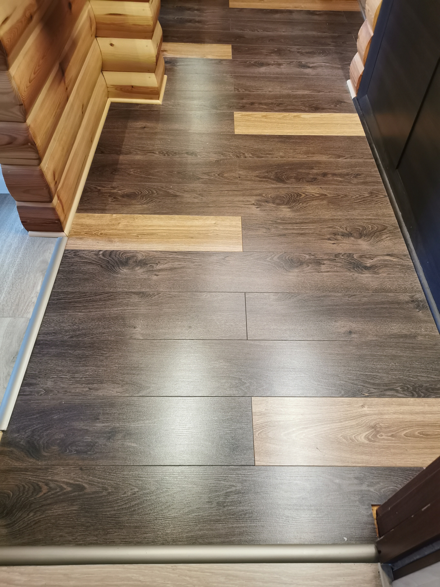 Ułożone panele podłogowe w odcieniach brązu i beżu, widoczne łączenia i listwy przypodłogowe, fragment drewnianej ściany z bali.