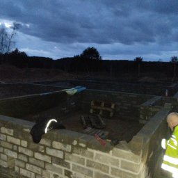 Tomasz Lewandowski - Murowanie fundamentów pod budynek w Stargardzie. Widoczny mur z cegieł, narzędzia i pracownik w odblaskowej kamizelce. Prace murarskie w toku, pochmurne niebo.