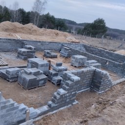Tomasz Lewandowski - Budowa fundamentów z bloczków betonowych na placu budowy. Częściowo wzniesione ściany fundamentowe, palety z bloczkami wewnątrz. W tle drzewa i hałda piasku.