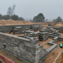 Tomasz Lewandowski - Wznoszenie fundamentów z bloczków betonowych na piaszczystym terenie. Widoczne narzędzia i materiały budowlane. Mglista pogoda w tle.