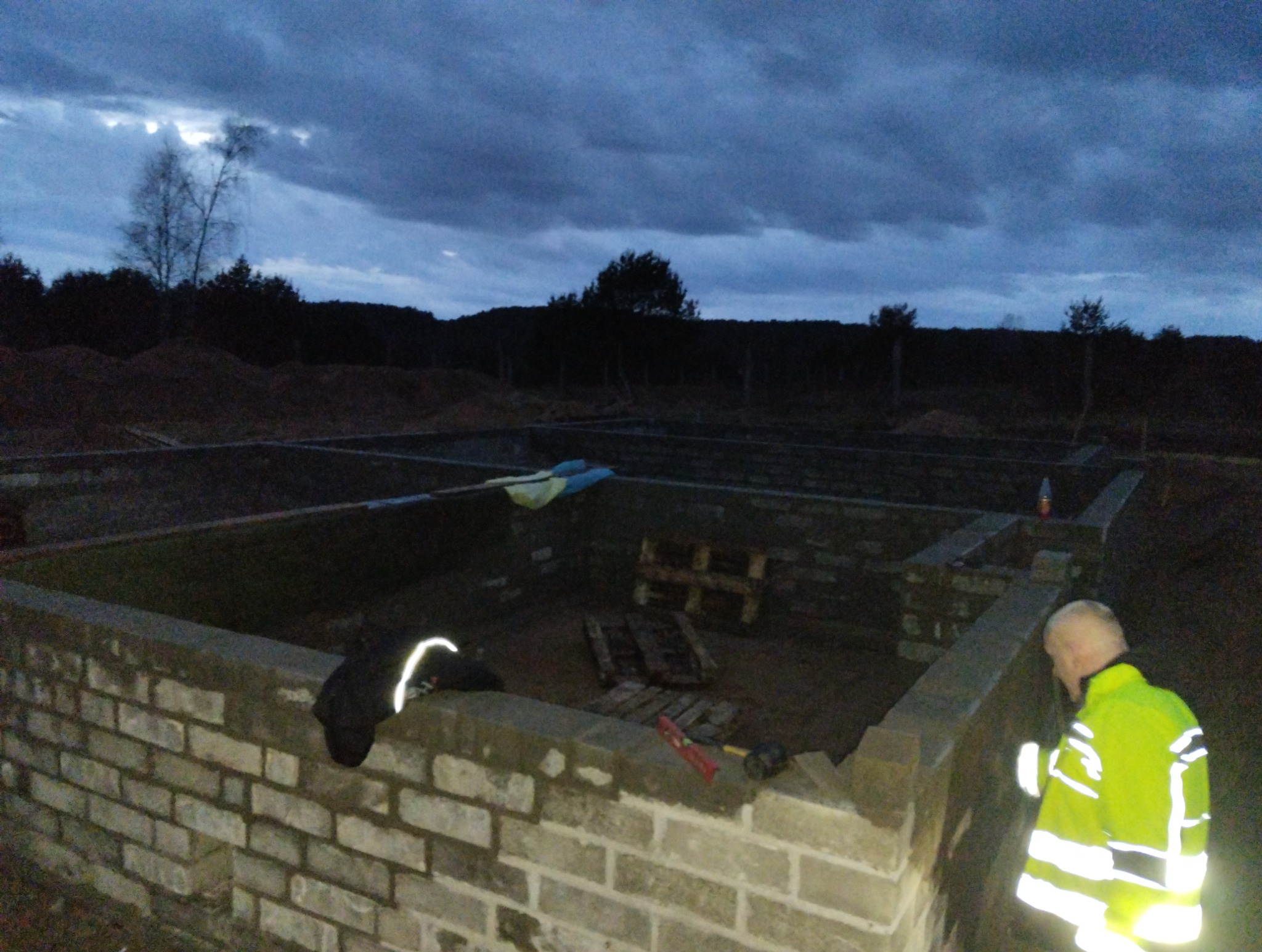 Murowanie fundamentów pod budynek w Stargardzie. Widoczny mur z cegieł, narzędzia i pracownik w odblaskowej kamizelce. Prace murarskie w toku, pochmurne niebo.