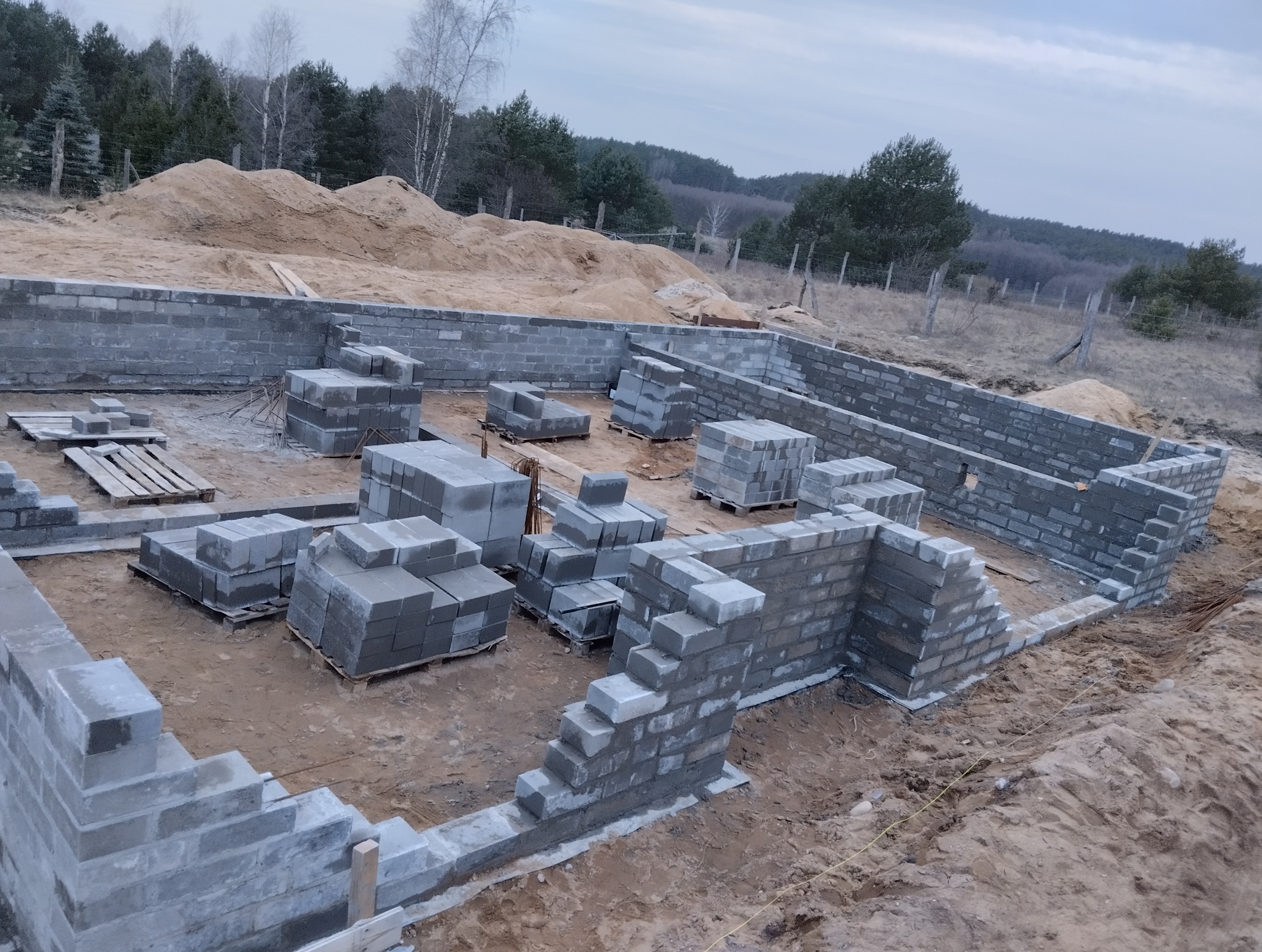 Budowa fundamentów z bloczków betonowych na placu budowy. Częściowo wzniesione ściany fundamentowe, palety z bloczkami wewnątrz. W tle drzewa i hałda piasku.