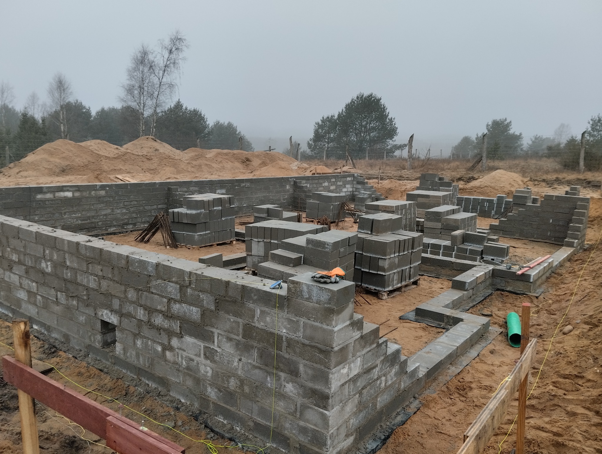 Wznoszenie fundamentów z bloczków betonowych na piaszczystym terenie. Widoczne narzędzia i materiały budowlane. Mglista pogoda w tle.