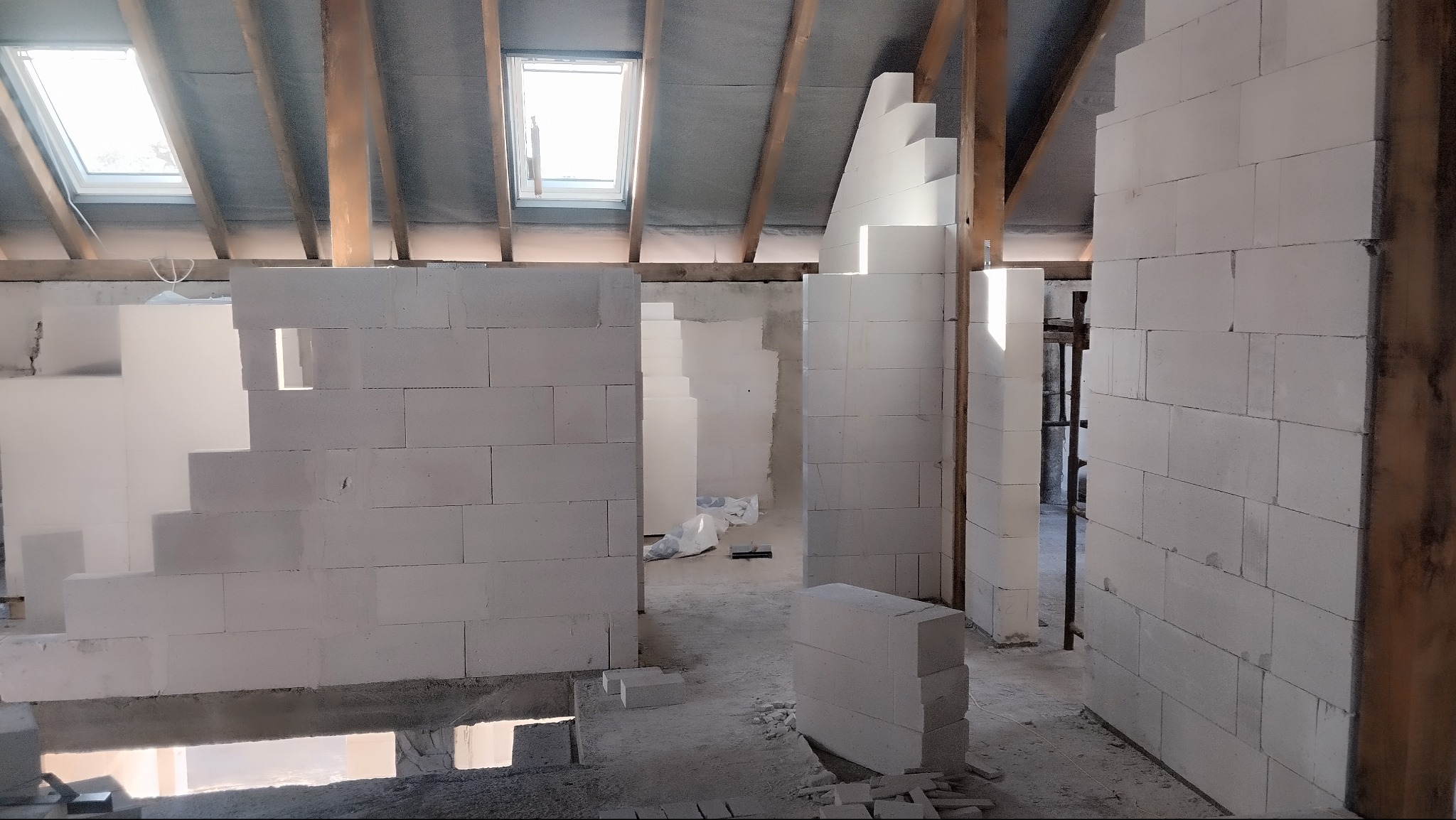 Wnętrze poddasza w trakcie budowy ścian działowych z białych bloczków, widoczne drewniane elementy konstrukcji dachu i okno dachowe. Surowy stan, etap murowania.