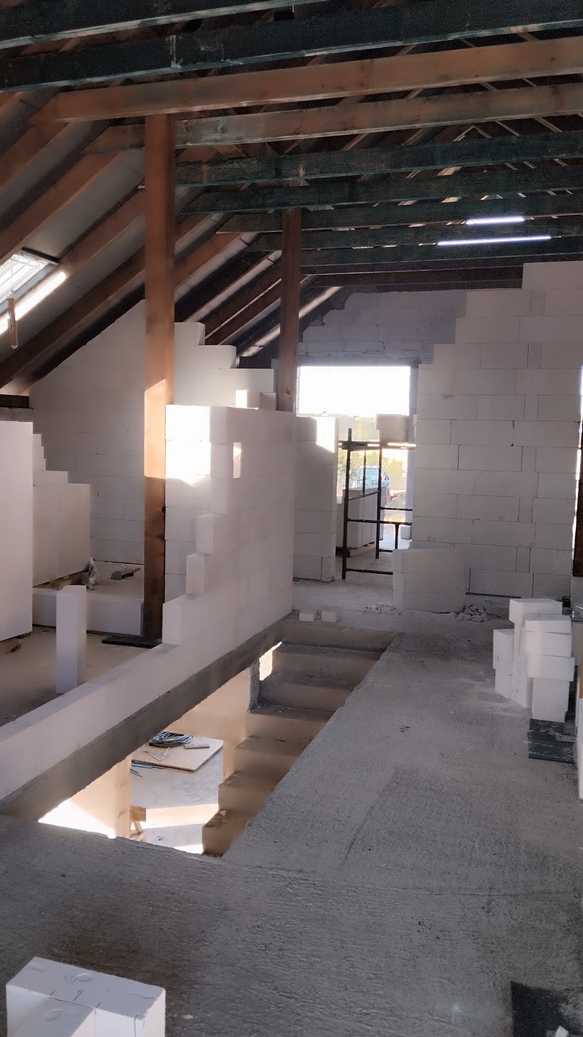 Wnętrze w trakcie budowy: surowy stan z widocznymi bloczkami, drewnianą konstrukcją dachu i wpadającym światłem słonecznym przez okno. Widoczne narzędzia i materiały budowlane.