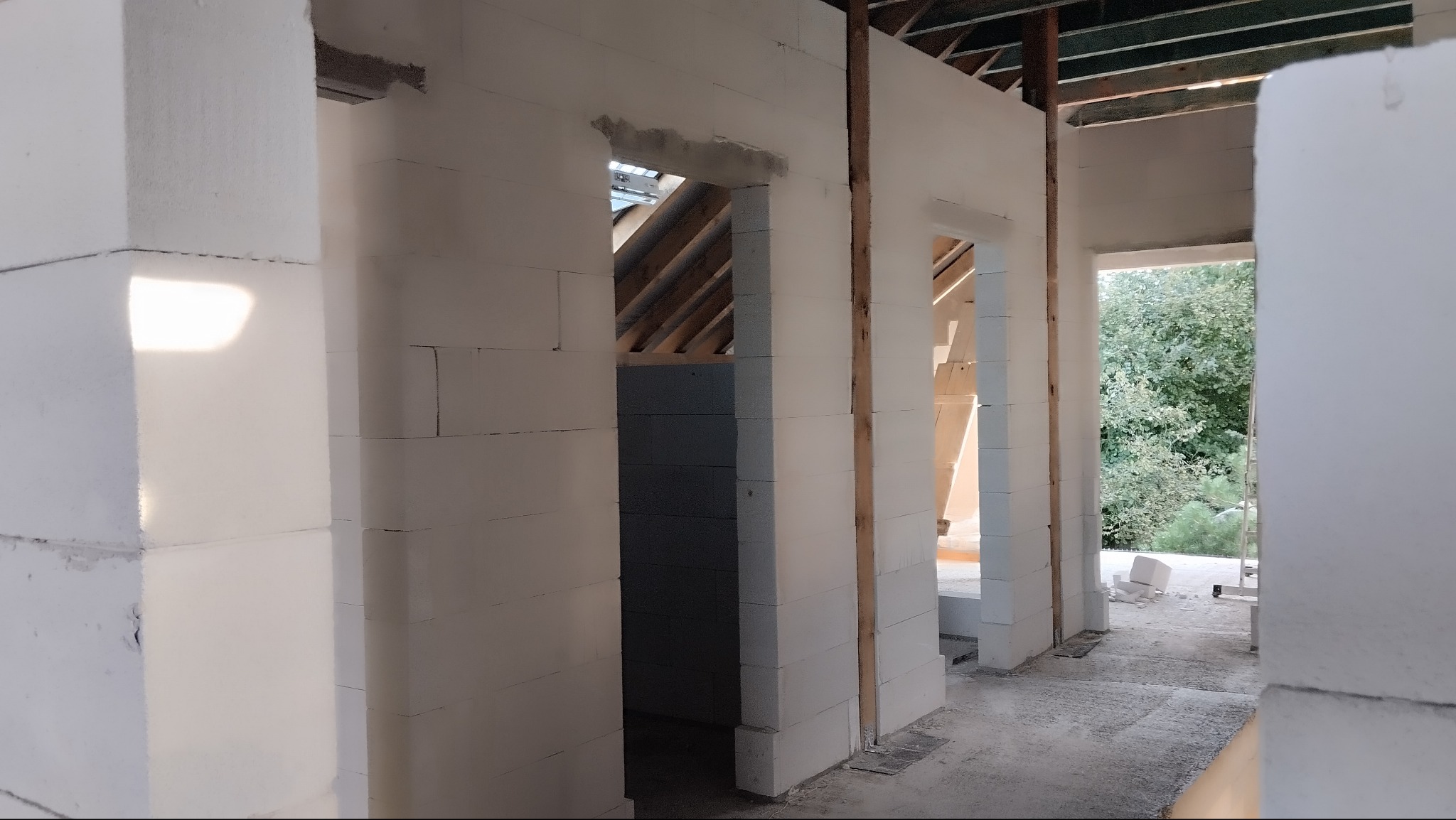 Wnętrze domu w trakcie budowy z białych bloczków, widoczne drewniane elementy konstrukcji dachu i otwory drzwiowe. Surowy stan, widok na zieleń przez okno.