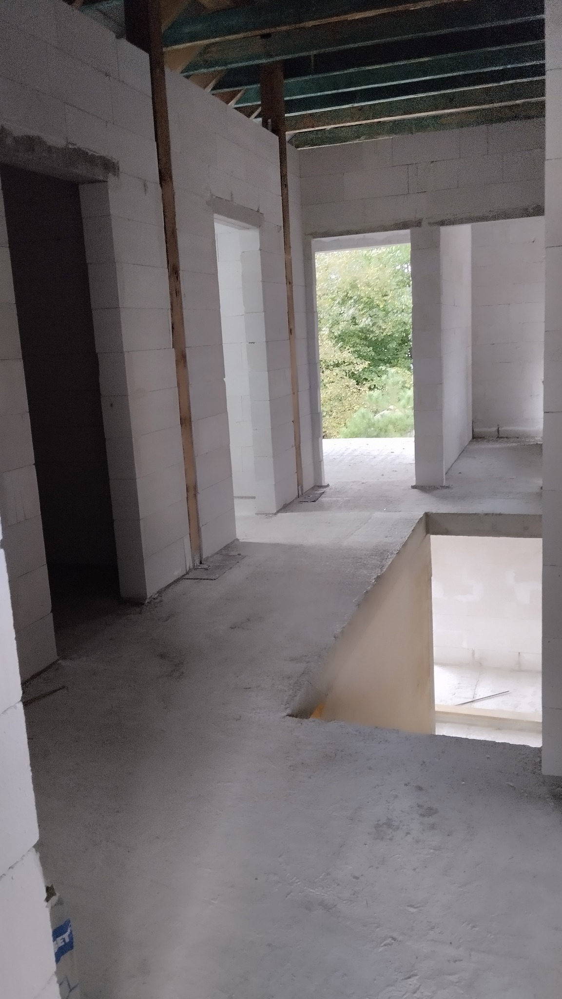 Surowy stan wnętrza budynku w trakcie budowy: białe ściany z betonu komórkowego, drewniane stemple, w tle widok na zieleń przez otwór okienny.
