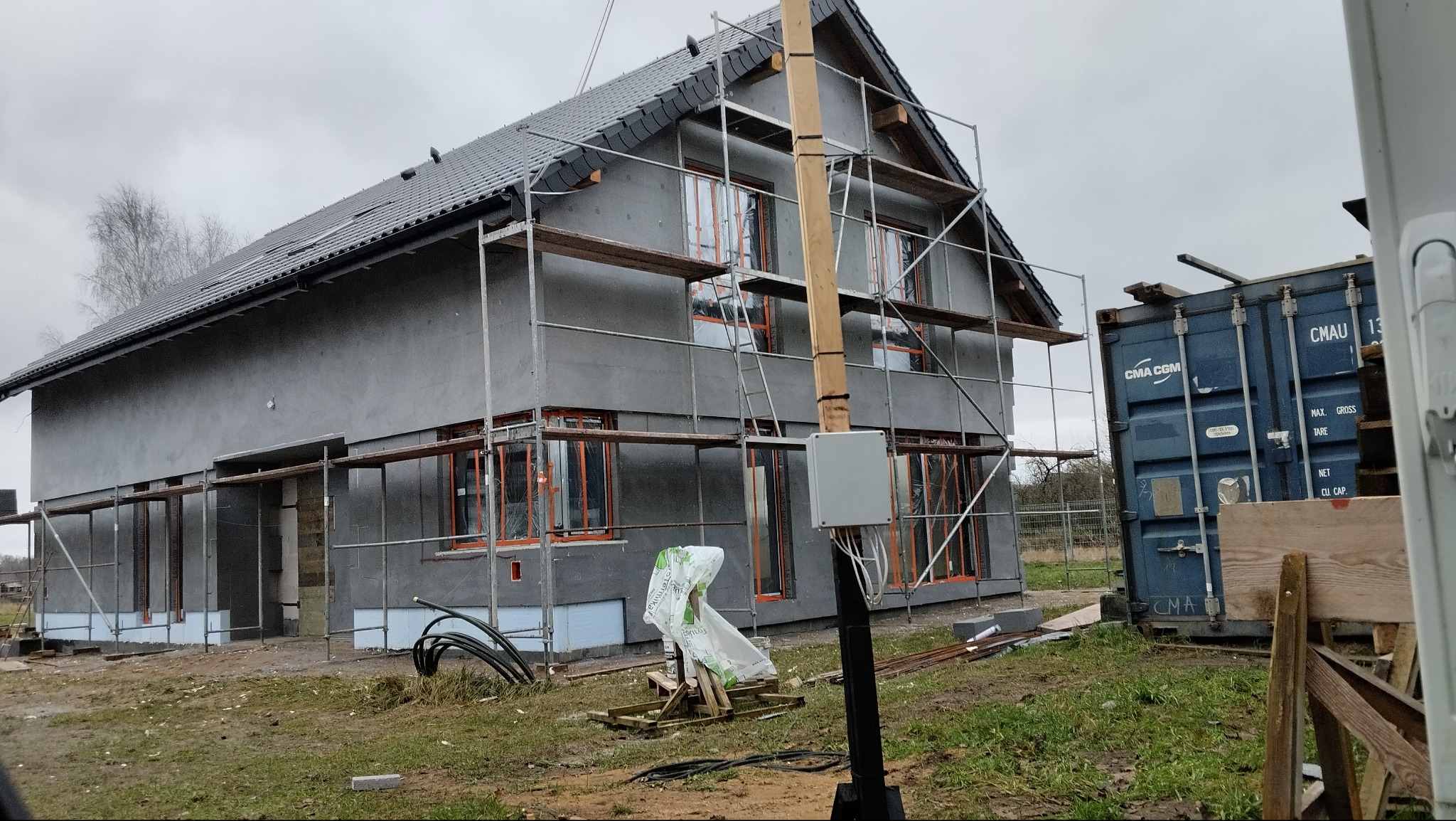Dom w trakcie ocieplania, z widocznymi rusztowaniami i kontenerem transportowym w tle. Szara elewacja, okna zabezpieczone folią, pochmurne niebo.