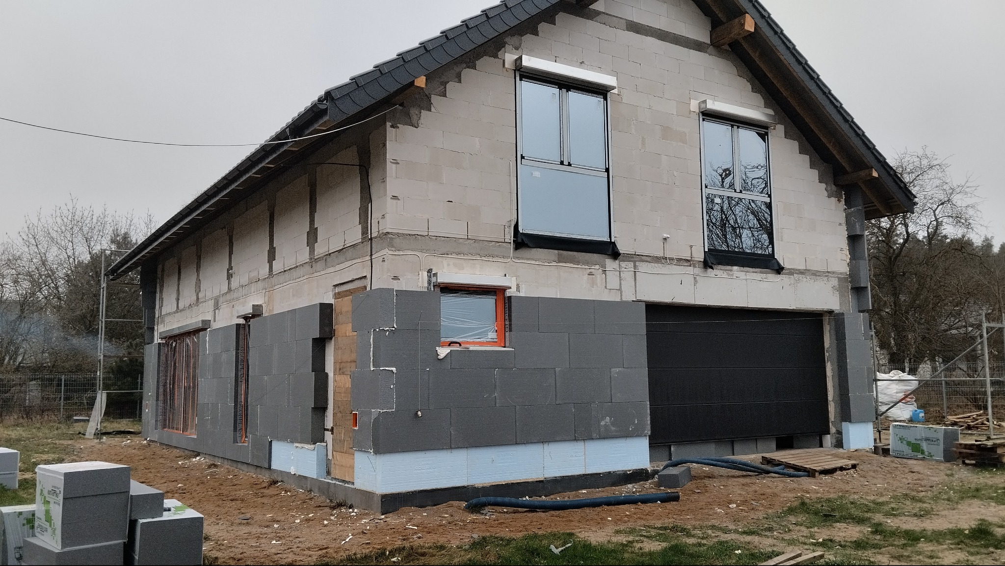 Budowa domu jednorodzinnego: ściany z bloczków, okna z roletami zewnętrznymi, częściowo ocieplone ściany szarym styropianem. Widoczne materiały budowlane.