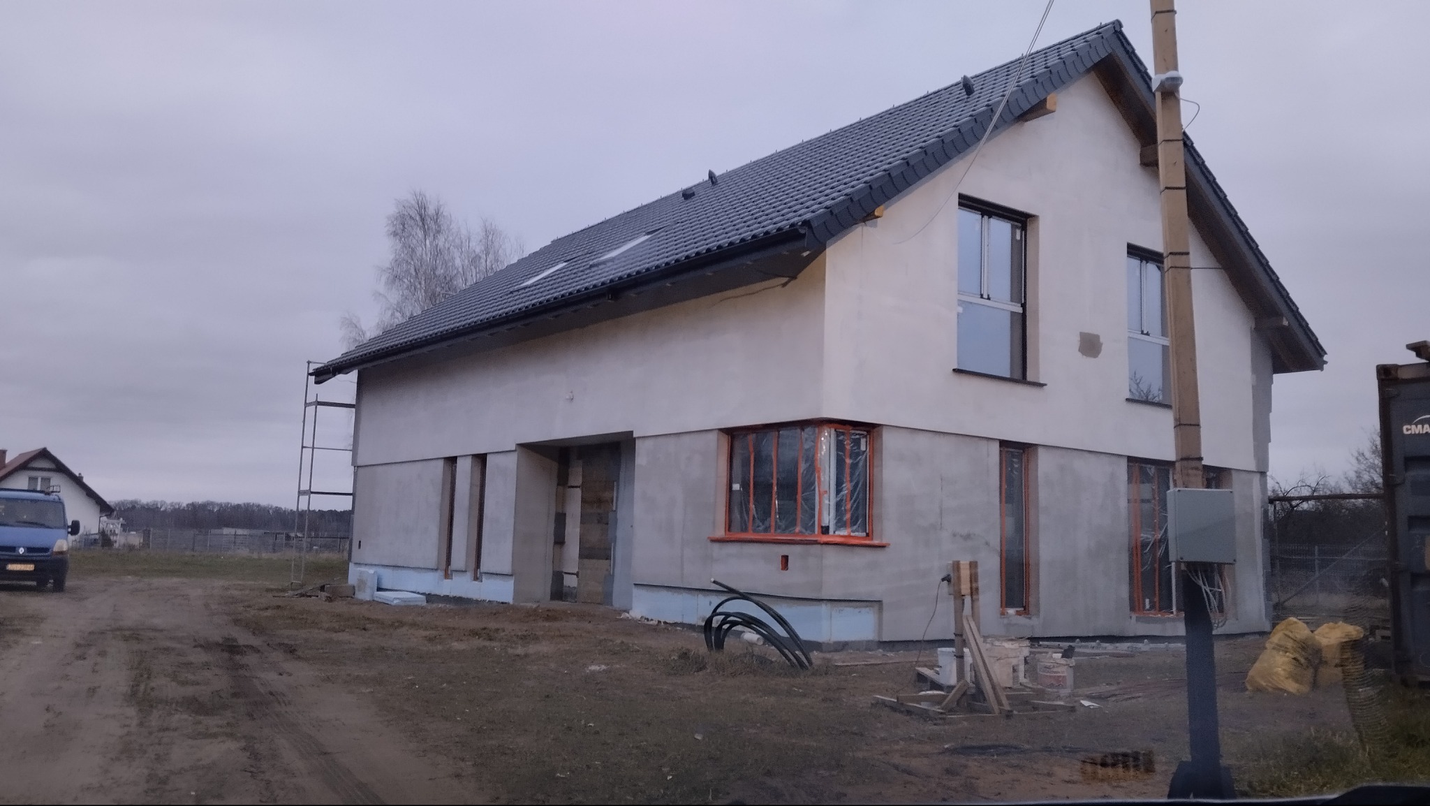 Surowy stan domu jednorodzinnego z szarą dachówką i oknami z pomarańczowymi ramami, częściowo otynkowany, z widocznymi materiałami budowlanymi na posesji.
