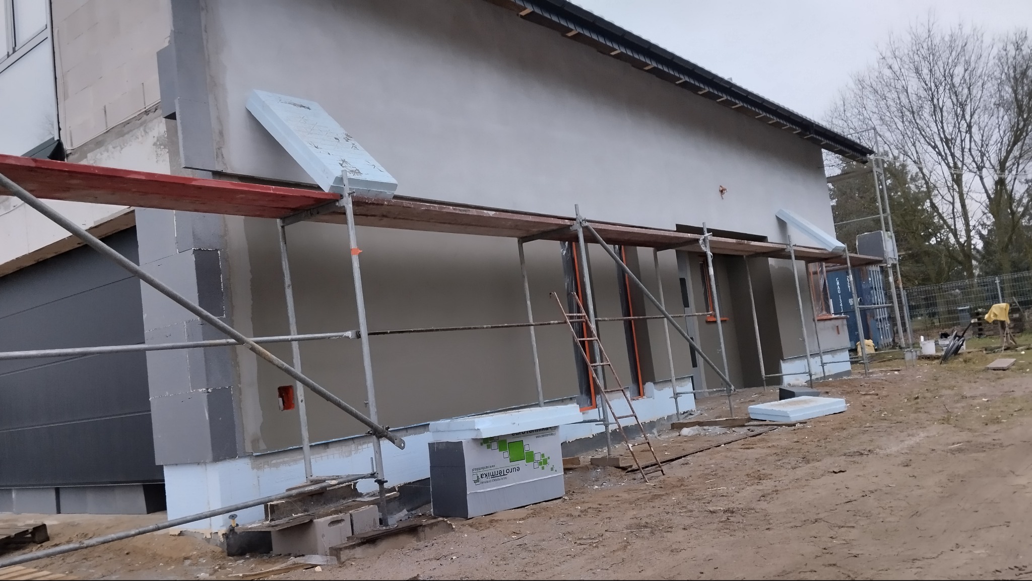 Budynek w trakcie ocieplania styropianem, widoczne rusztowania i materiały budowlane. Świeżo nałożony tynk na elewacji. Prace wykończeniowe w toku.