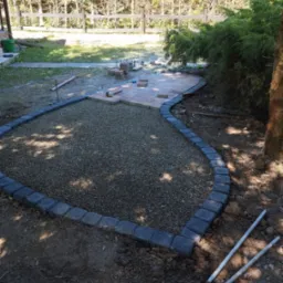 Układanie kostki brukowej na ścieżce ogrodowej, widoczne obrzeża z kostki, podsypka żwirowa i częściowo ułożona nawierzchnia z kostki brukowej w odcieniach beżu i brązu.