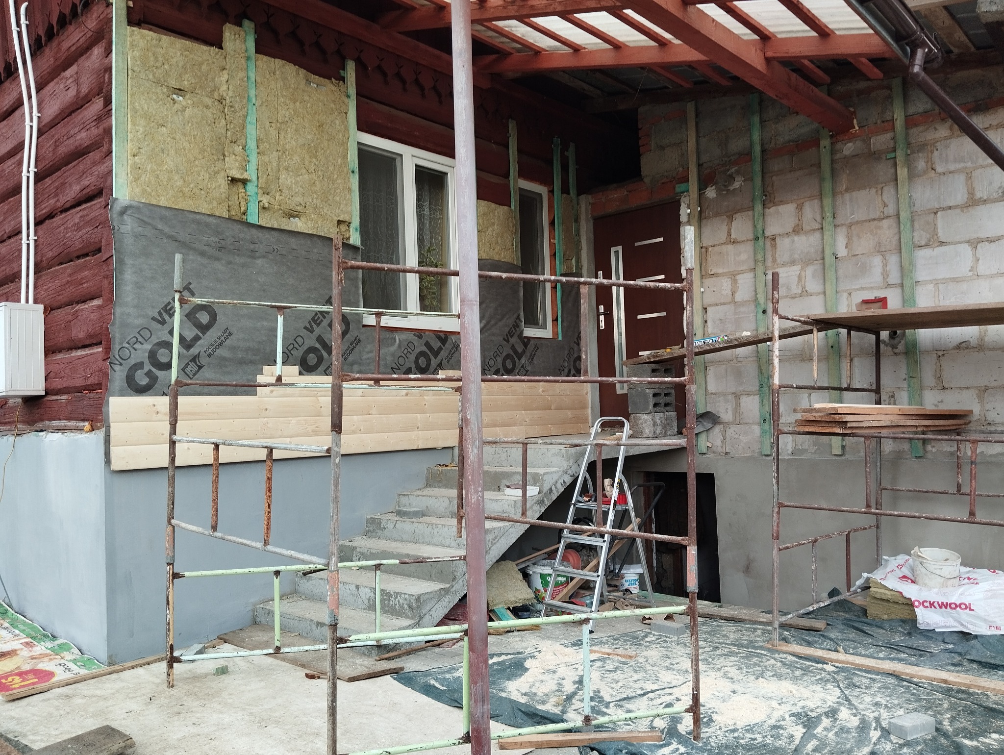 Ocieplanie drewnianego domu wełną mineralną i montaż paneli elewacyjnych, widoczne rusztowania i materiały budowlane.