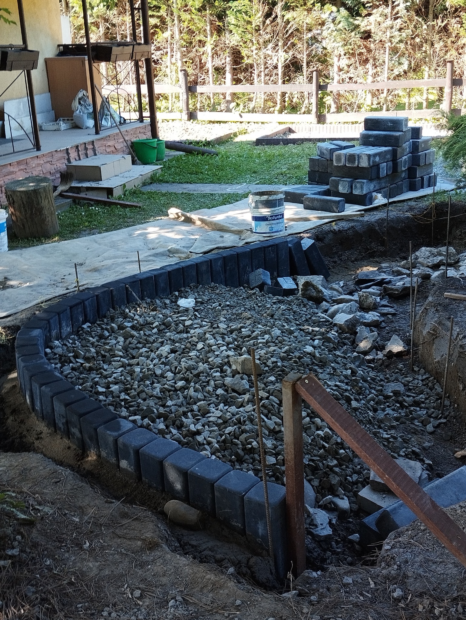 Przygotowanie podłoża pod ścieżkę z kostki: obrzeże z kostki, wysypane kamieniami, widoczne narzędzia i materiały budowlane w tle.