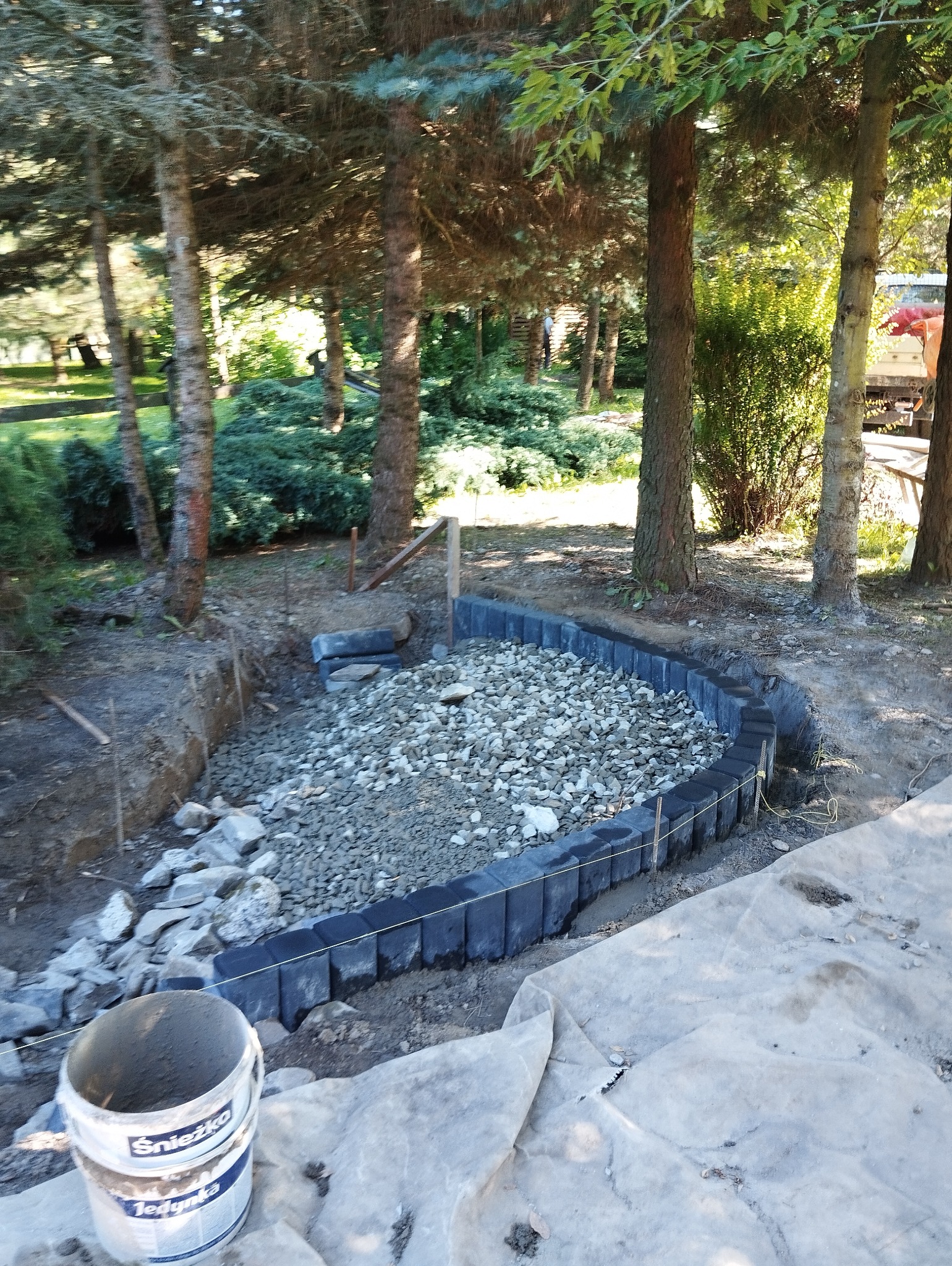 Przygotowanie podłoża pod ścieżkę w ogrodzie: wykop, obrzeża z kostki, wysypany żwir, widoczny sznurek wyznaczający linię, wiadro z zaprawą Snieżka Jedynka w rogu.