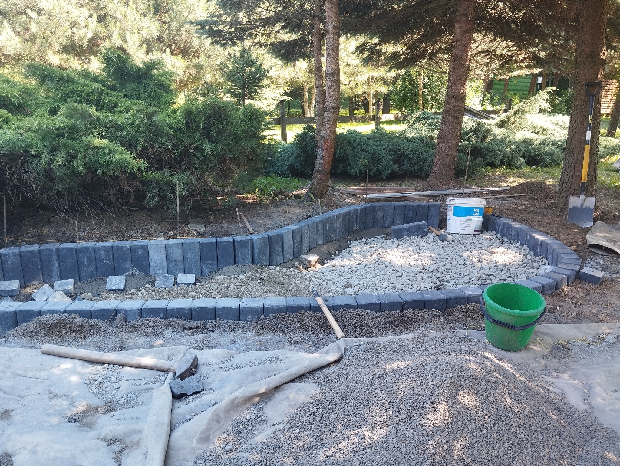 Krawężnik z ciemnej kostki obramowujący teren wysypany drobnym żwirem, widoczny biały pojemnik z klejem, zielone wiadro i narzędzia ogrodnicze, w tle drzewa iglaste.