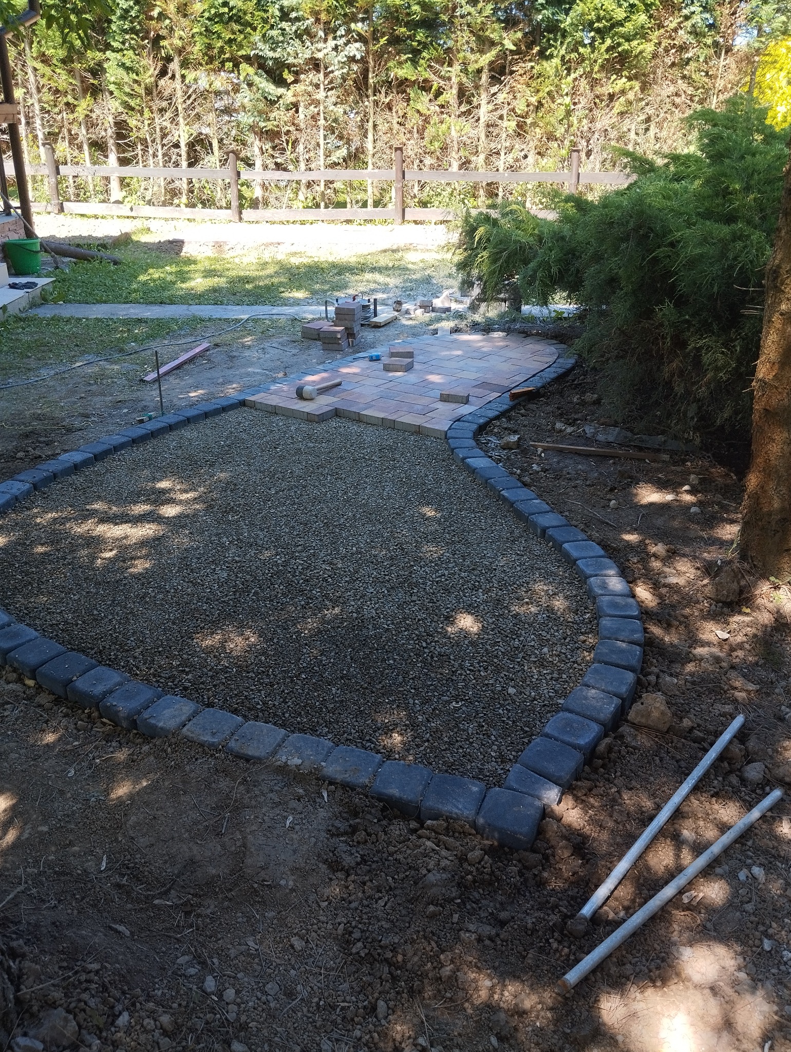 Układanie kostki brukowej na ścieżce ogrodowej, widoczne obrzeża z kostki, podsypka żwirowa i częściowo ułożona nawierzchnia z kostki brukowej w odcieniach beżu i brązu.