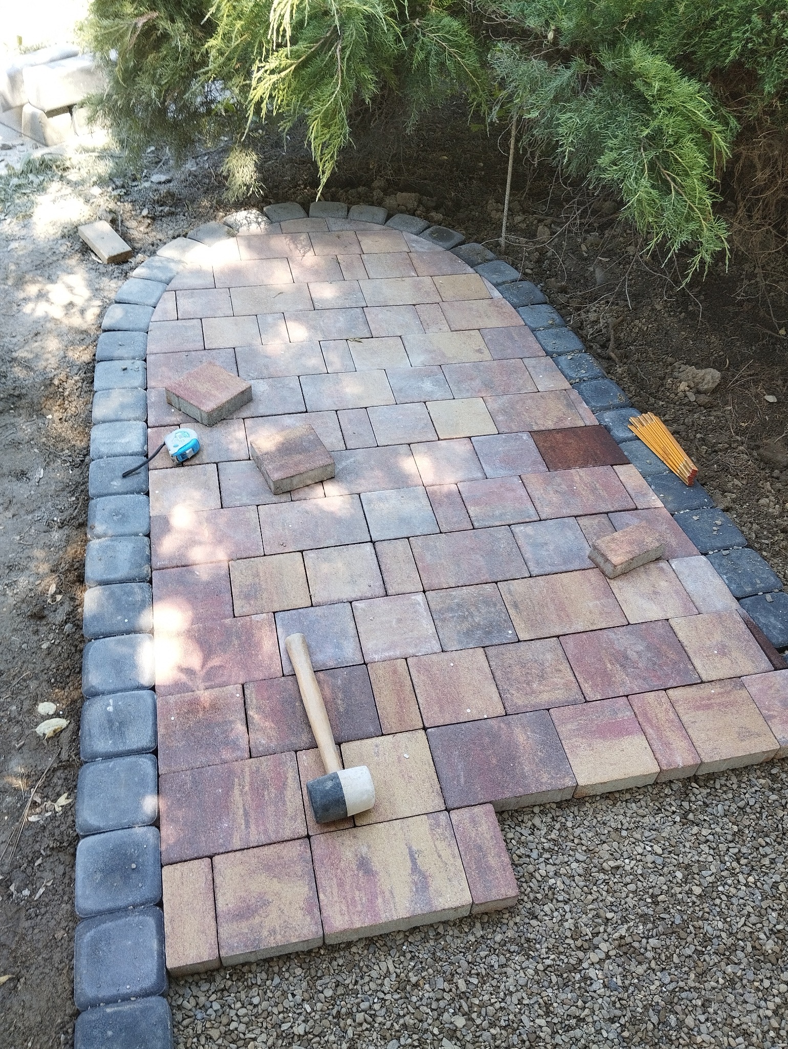 Fragment ścieżki z kostki brukowej w trakcie układania, widoczny gumowy młotek, miarka zwijana i składana, krawężniki z ciemnej kostki, podłoże z drobnego żwiru, w tle zieleń.