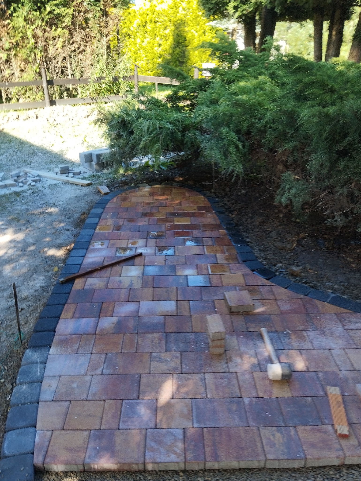 Świeżo ułożony, zakrzywiony chodnik z kostki brukowej w odcieniach brązu i czerni, otoczony zielenią, z widocznymi narzędziami pracy i stosem kostek, częściowo mokry od deszczu.