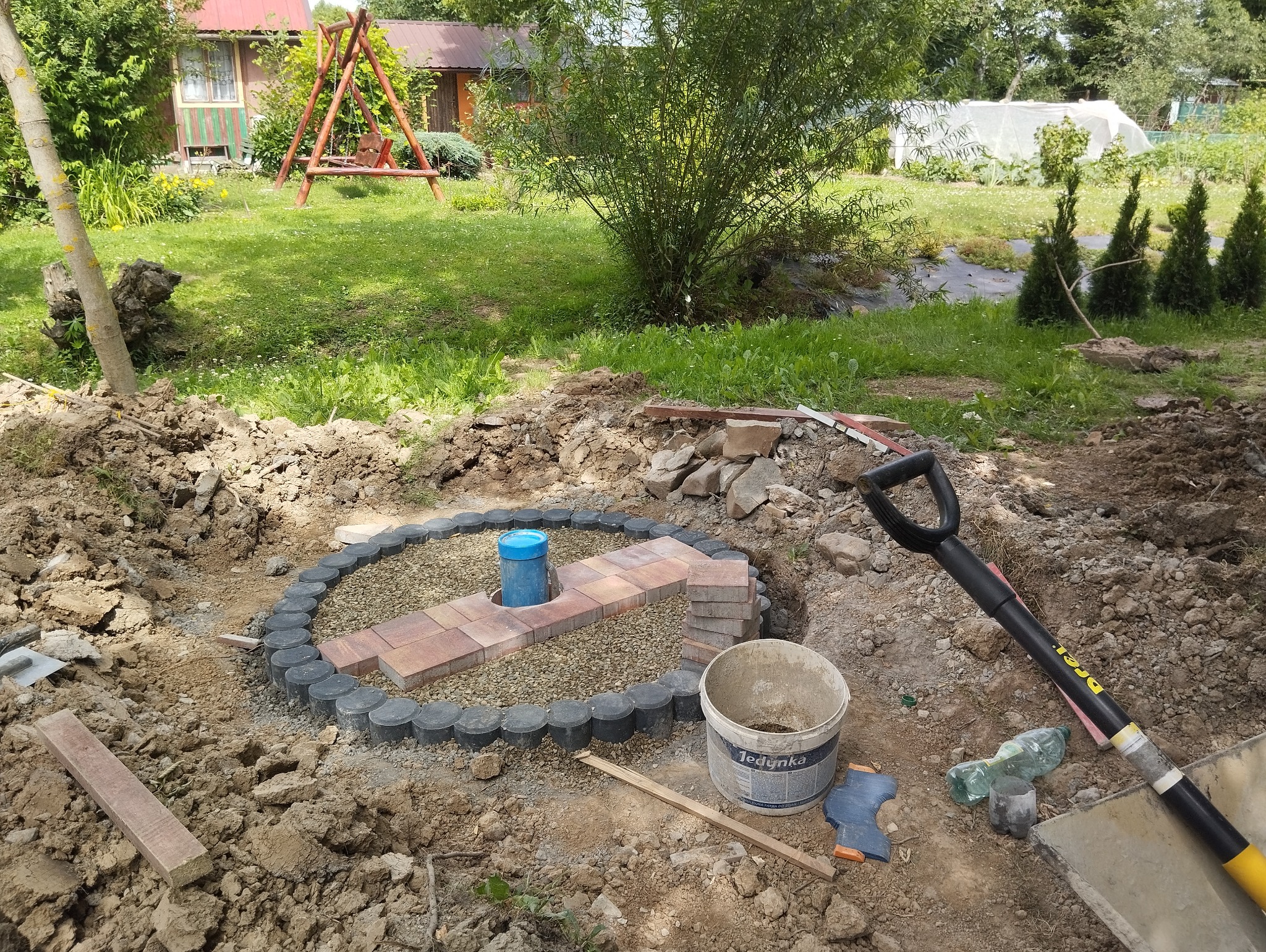Okrągła rabata w trakcie tworzenia: obrzeże z betonowych elementów, wypełnienie żwirowe, ścieżka z kostki, rura PCV w centrum, narzędzia i materiały budowlane wokół, ogród w tle.