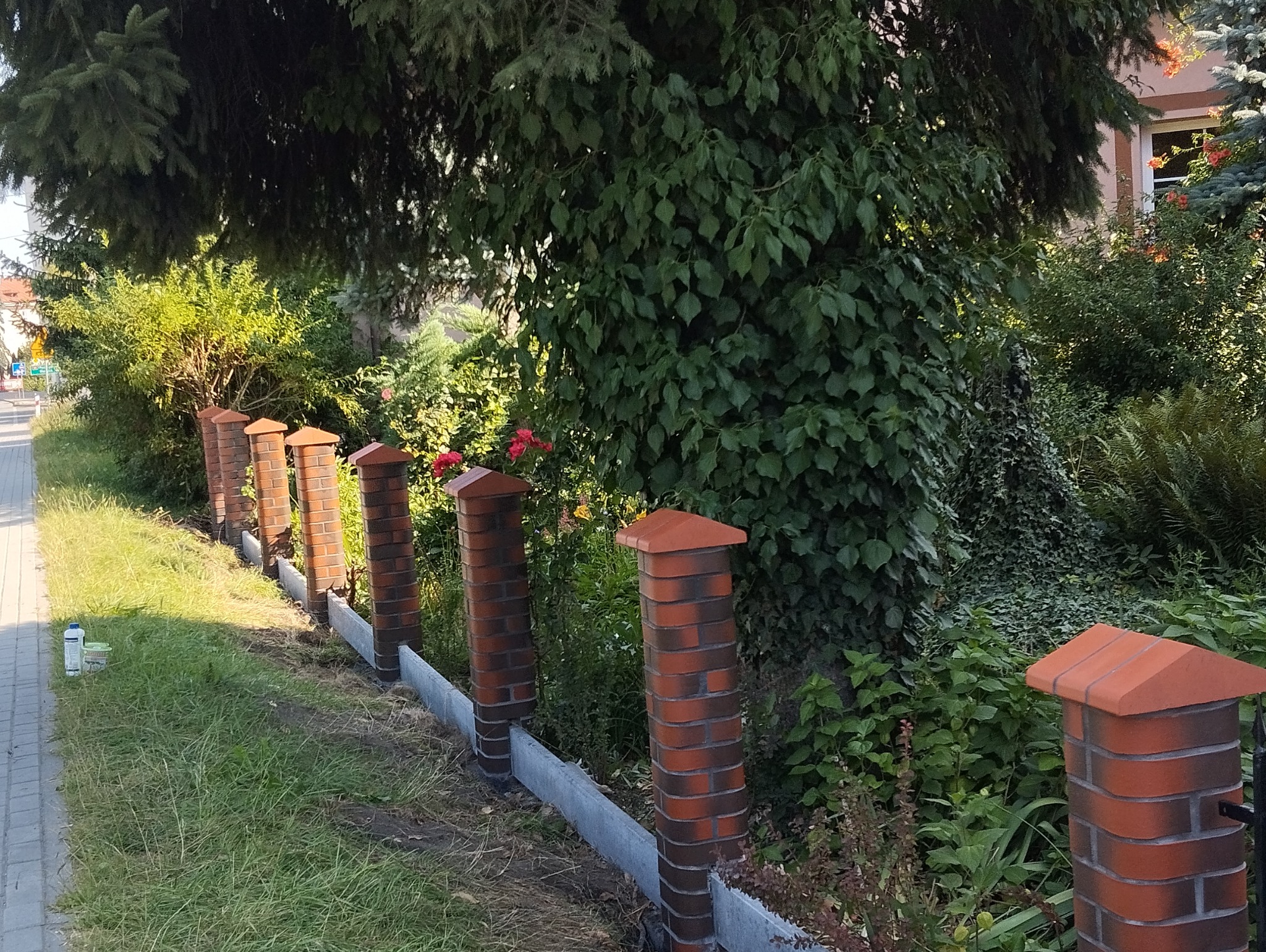 Fragment nowo wzniesionego ogrodzenia z ceglanych słupków i betonowej podmurówki, widoczny wzdłuż chodnika, z bujną zielenią w tle.
