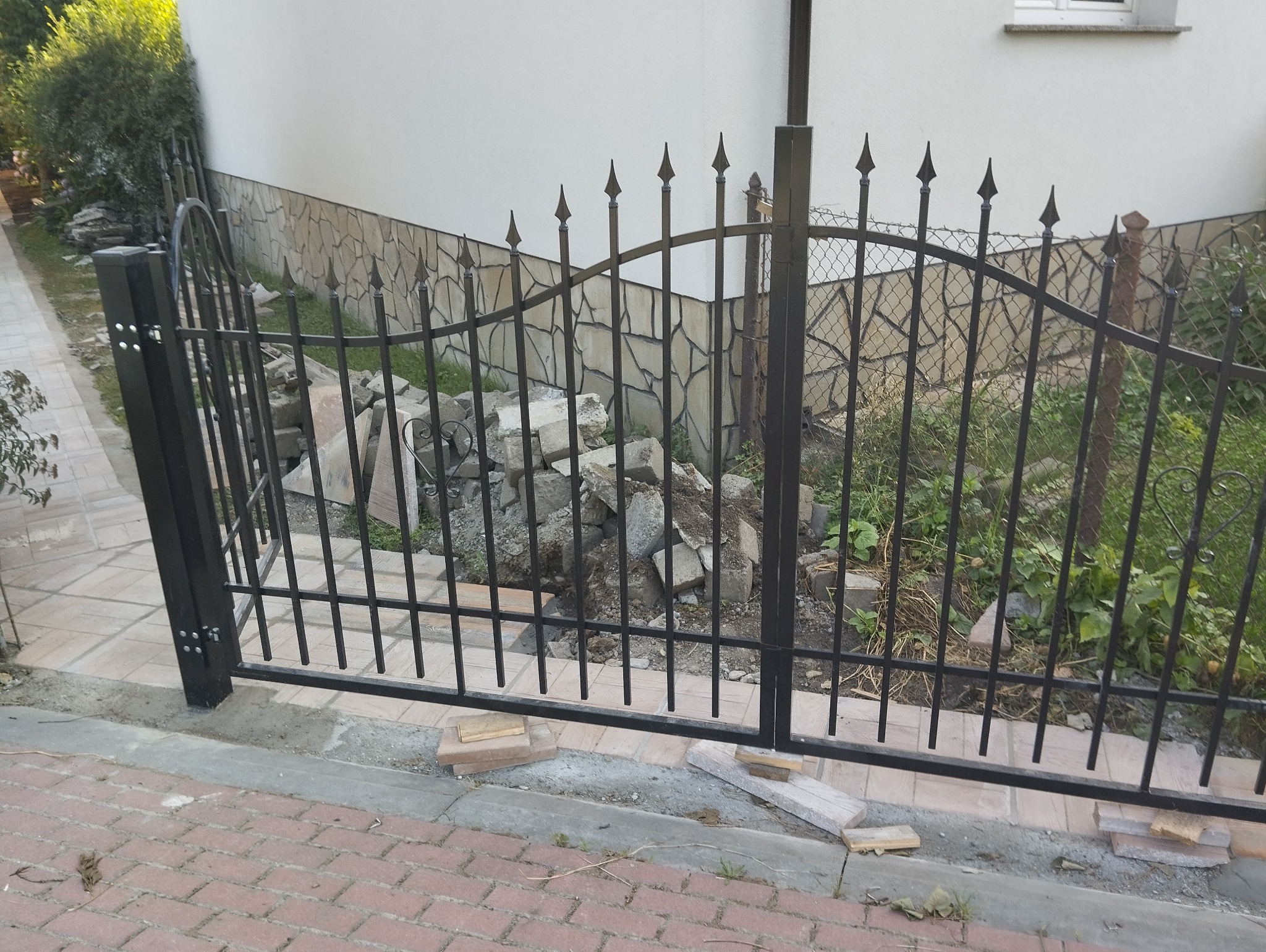 Czarna, metalowa brama z ostrymi zakończeniami, widok na stertę gruzu i roślinność w tle, częściowo zamontowana na ceglanym chodniku.
