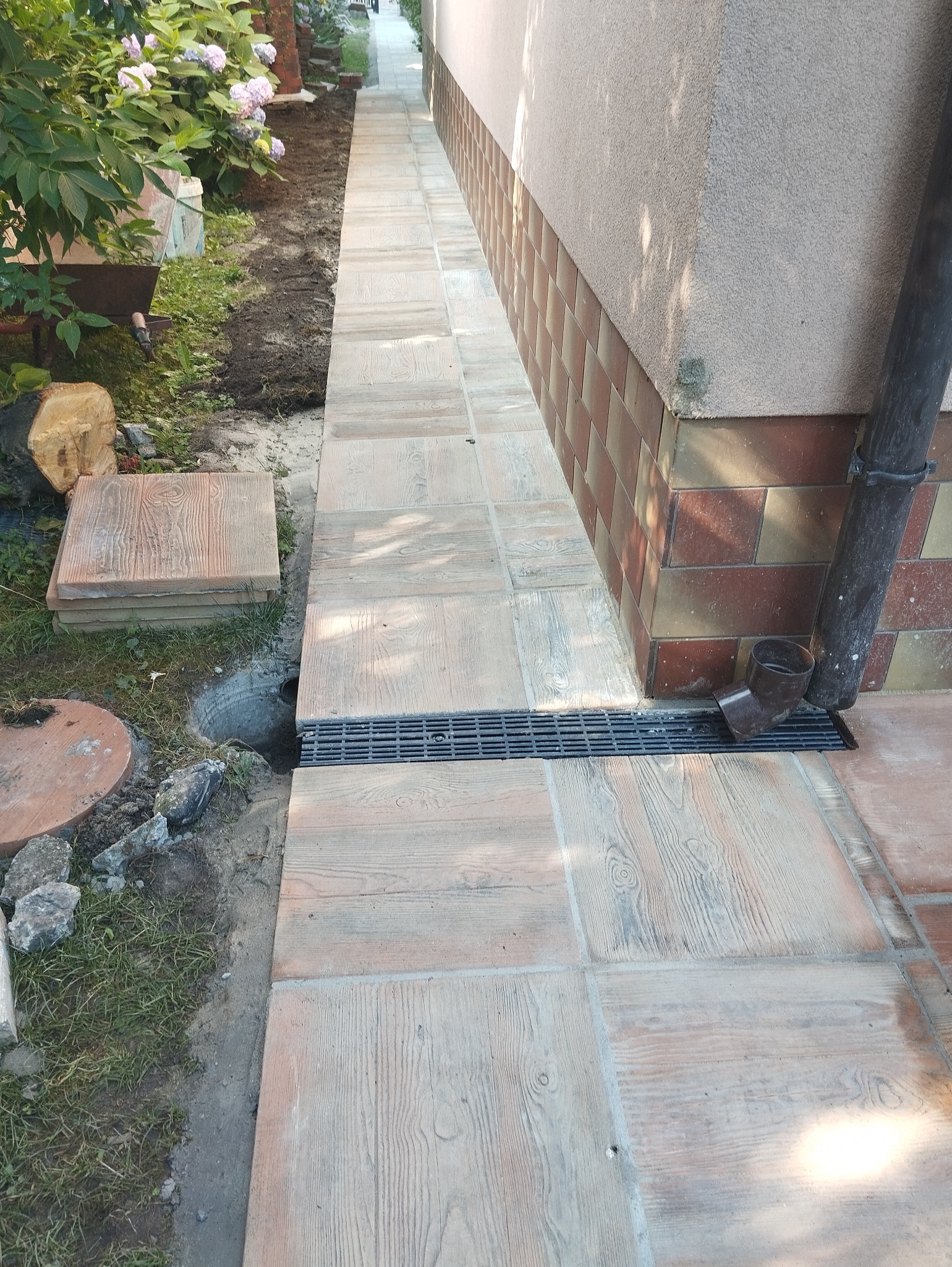 Ułożona ścieżka z płyt imitujących drewno, z odpływem liniowym przy ścianie budynku z okładziną z cegieł, obok wykop i stos płyt do ułożenia.