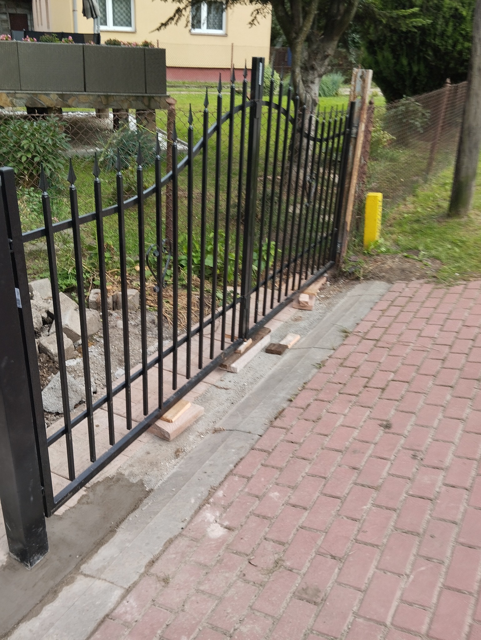 Montaż czarnego, metalowego ogrodzenia z ostrymi zakończeniami, widoczne cegły podtrzymujące konstrukcję, świeży beton przy słupku oraz czerwona kostka brukowa.