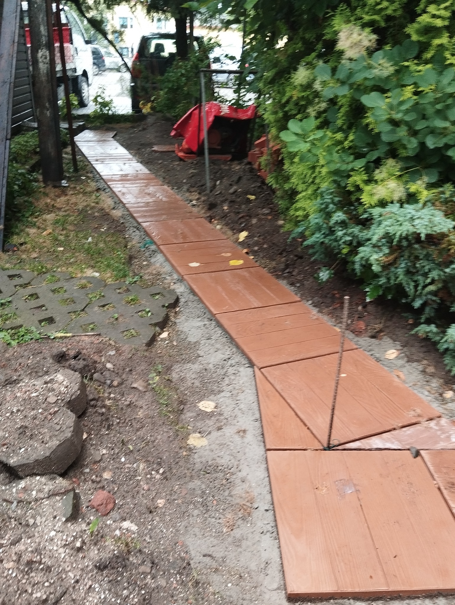 Świeżo ułożony chodnik z brązowych płyt imitujących drewno, prowadzący przez ogród, z widocznymi elementami mocującymi i fragmentami starej nawierzchni obok.