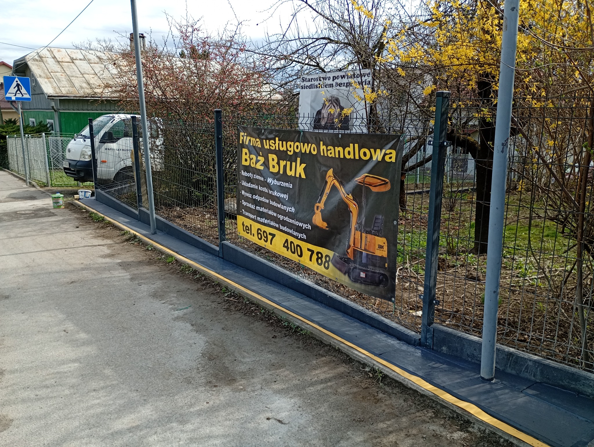 Baner reklamowy firmy Baż Bruk z ilustracją koparki umieszczony na ogrodzeniu z siatki, w tle budynek i znak drogowy.