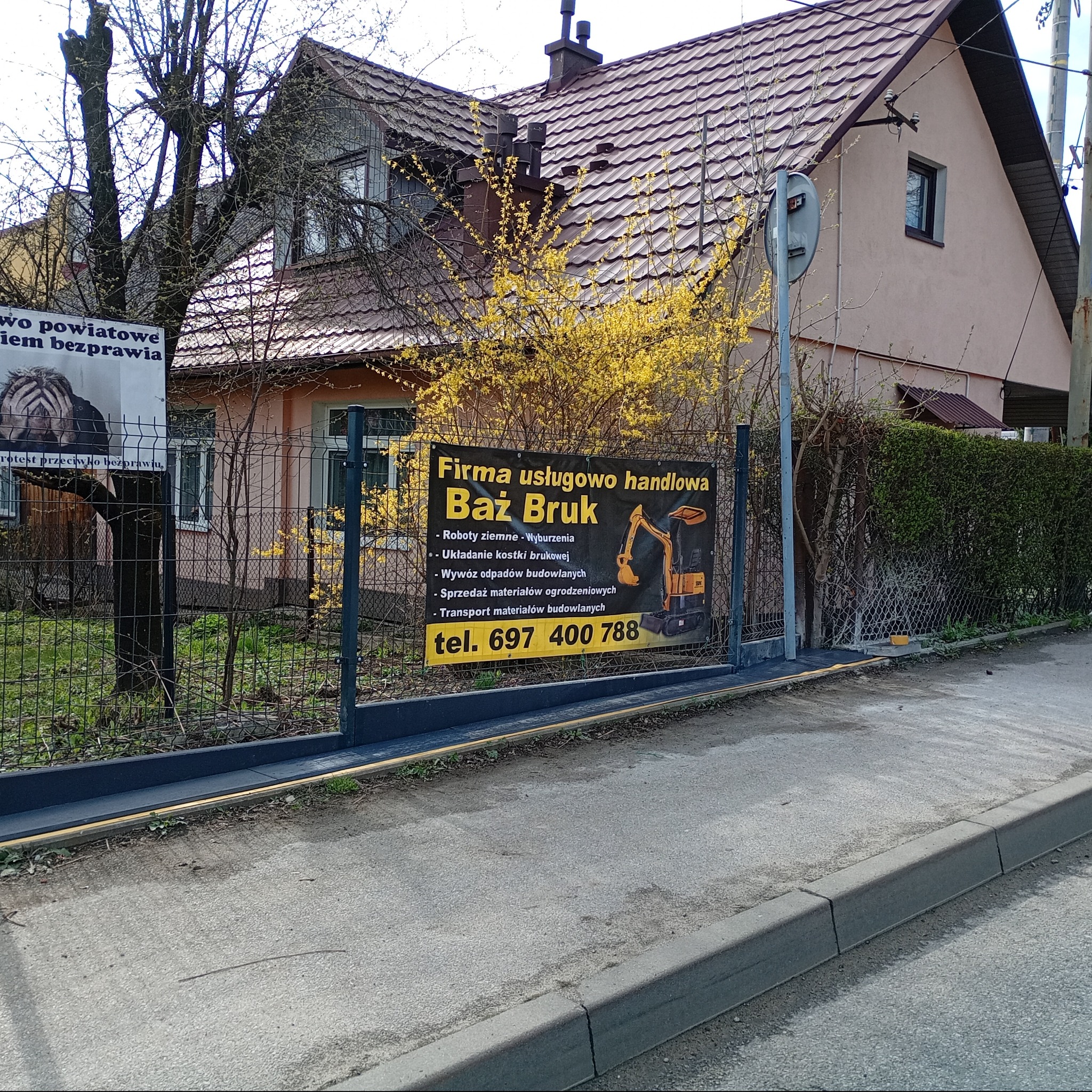 Ogłoszenie firmy Baż Bruk na ogrodzeniu posesji, oferującej roboty ziemne, wyburzenia, układanie kostki, wywóz odpadów i transport materiałów budowlanych.