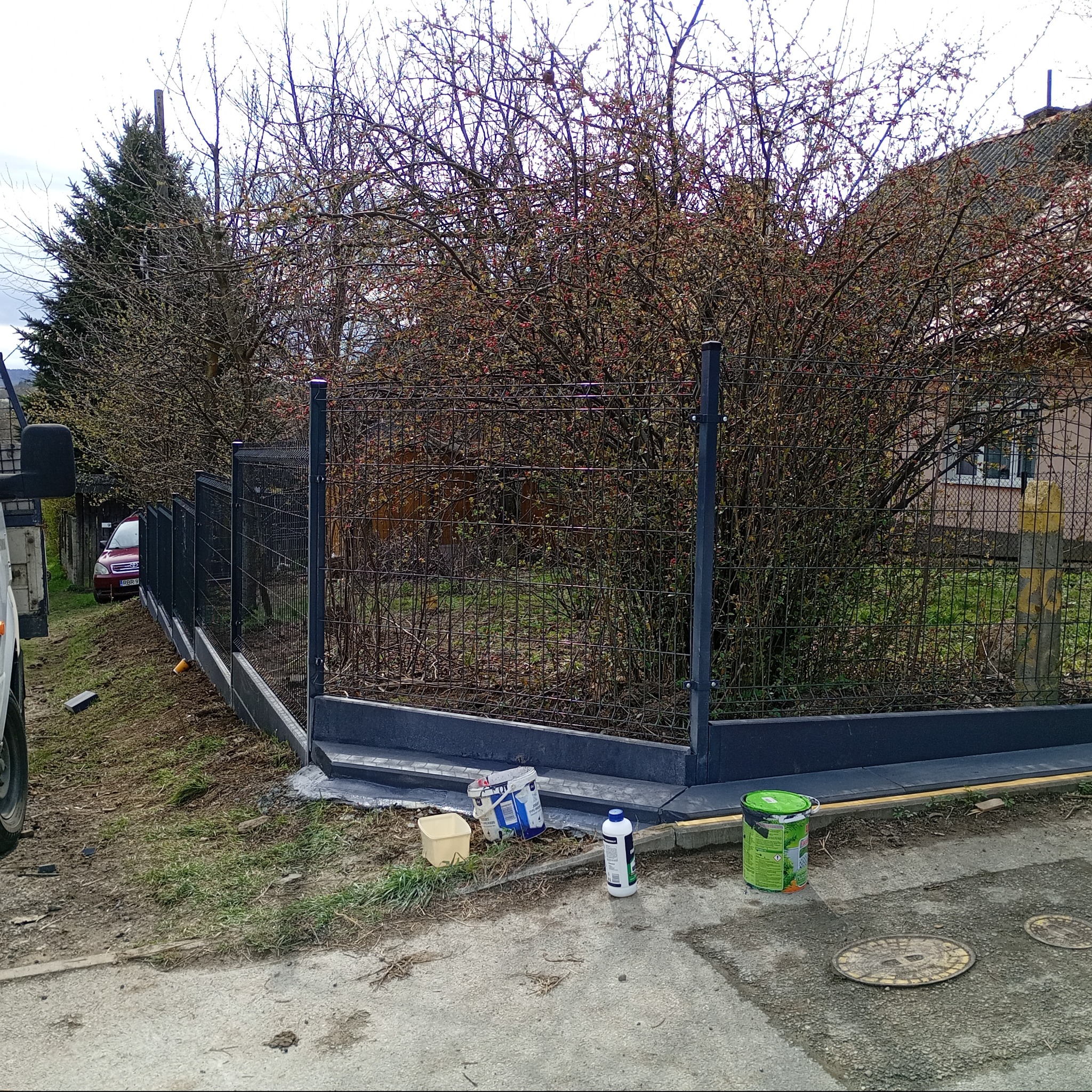 Nowo postawione, ciemnoszare ogrodzenie panelowe na betonowej podmurówce, widoczne puszki po farbie i impregnacie, w tle krzewy i dom.