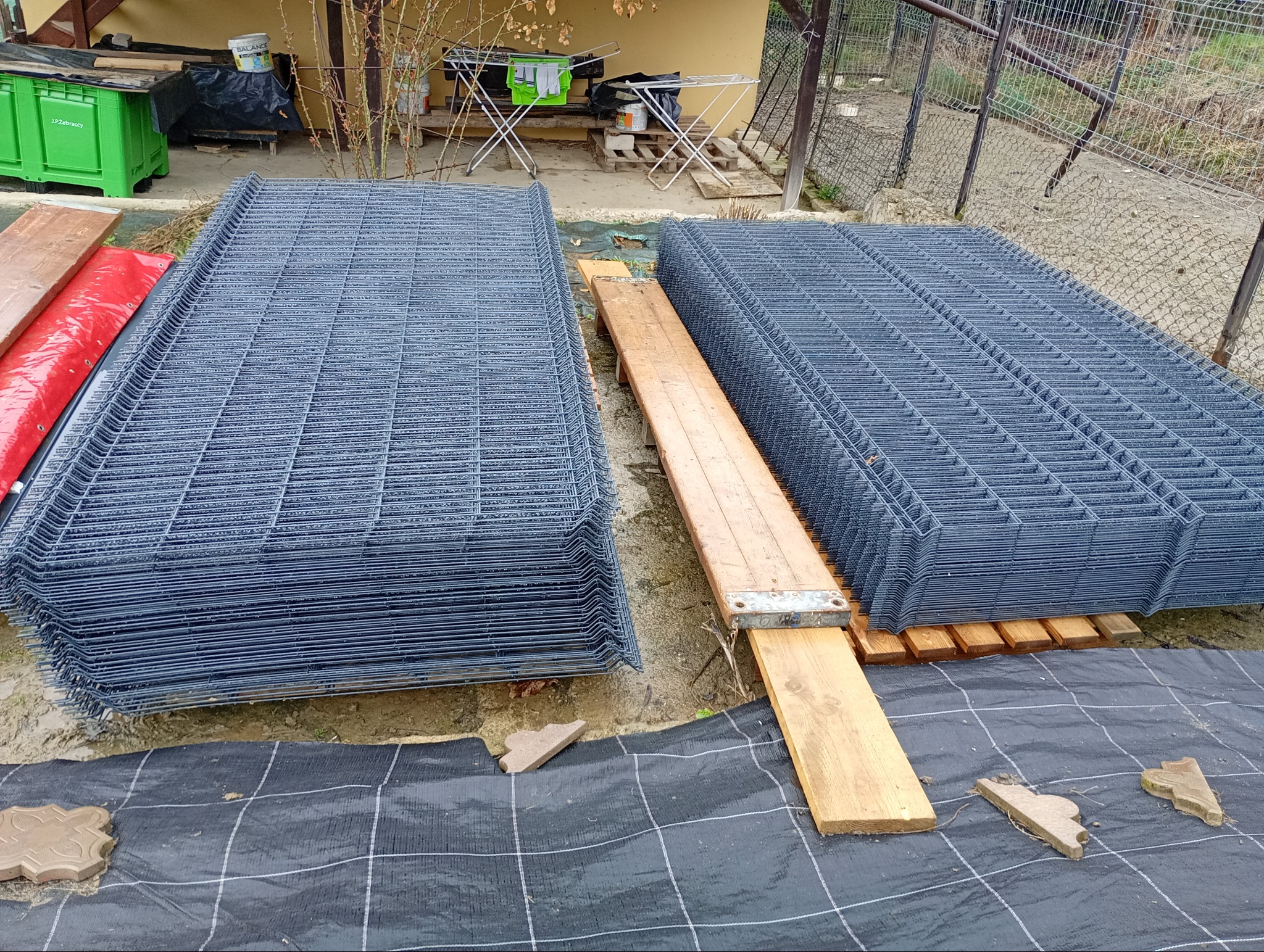 Stosy paneli ogrodzeniowych zgrzewanych, zabezpieczonych drewnianymi belkami, leżące na czarnej geowłókninie z kostką brukową w tle, przygotowane do transportu lub montażu.