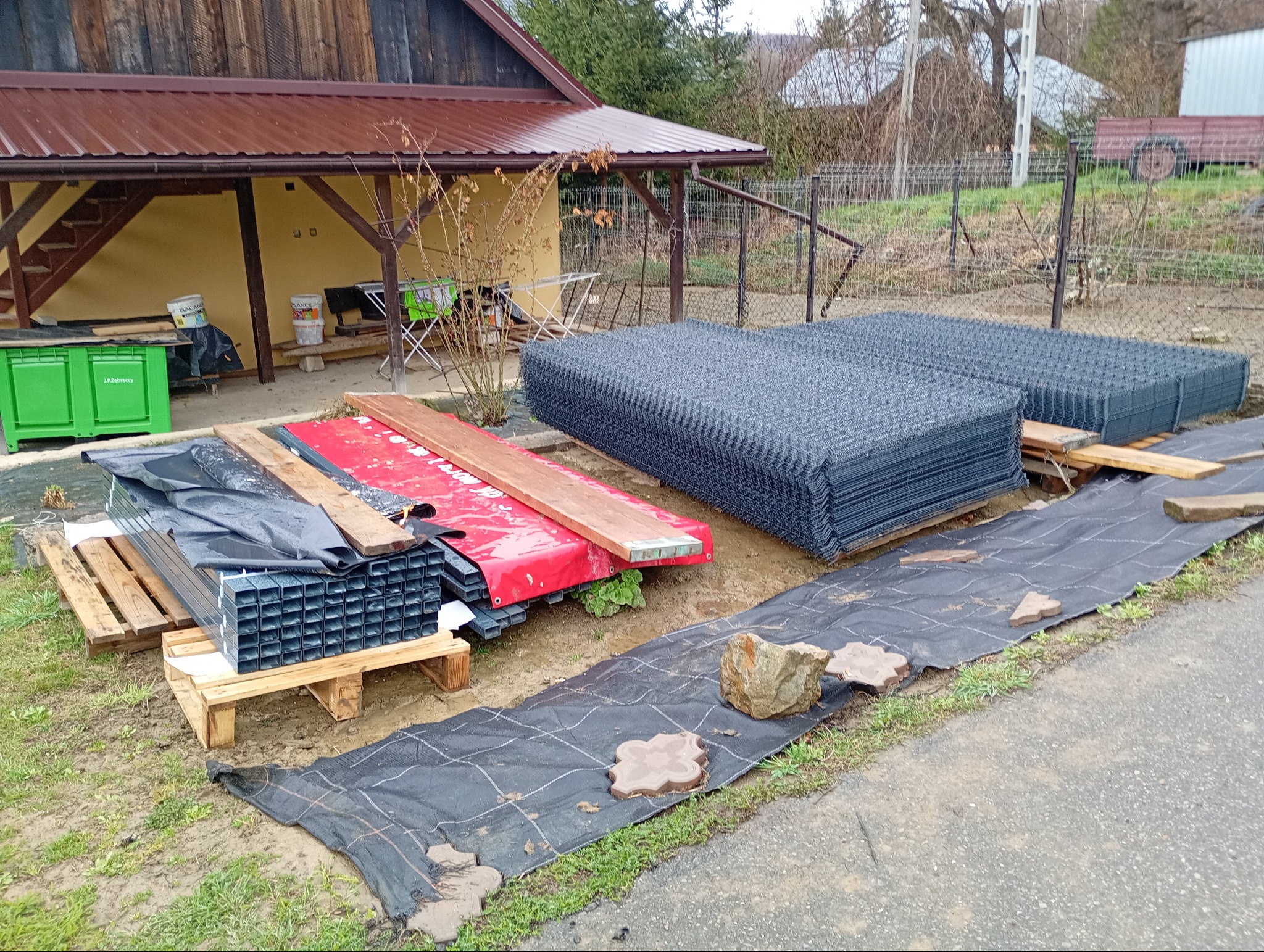 Przygotowane materiały do budowy ogrodzenia: panele ogrodzeniowe, słupki profilowe, drewniane elementy, geowłóknina oraz kostka brukowa na posesji przed domem.