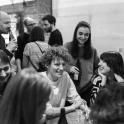 Czarno-białe zdjęcie grupy uśmiechniętych osób siedzących przy stole z kieliszkami i butelkami, w tle mur z cegły i plakat.