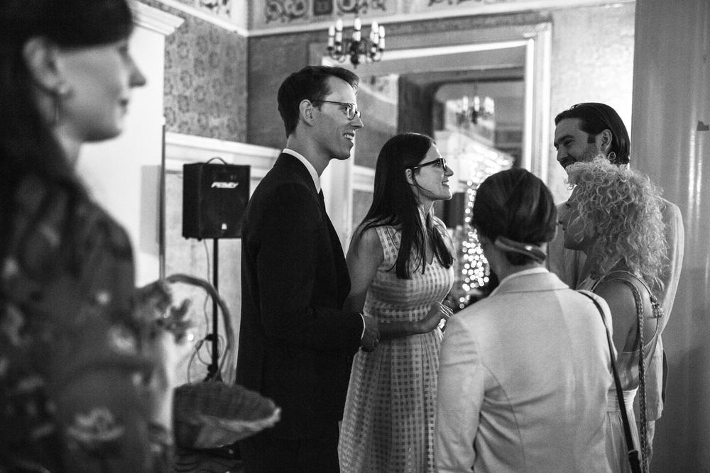 Czarno-białe zdjęcie przedstawia grupę osób w eleganckim wnętrzu z żyrandolami, prawdopodobnie podczas przyjęcia weselnego; widoczny mężczyzna w garniturze i kobieta w sukience w kratę rozmawiają...