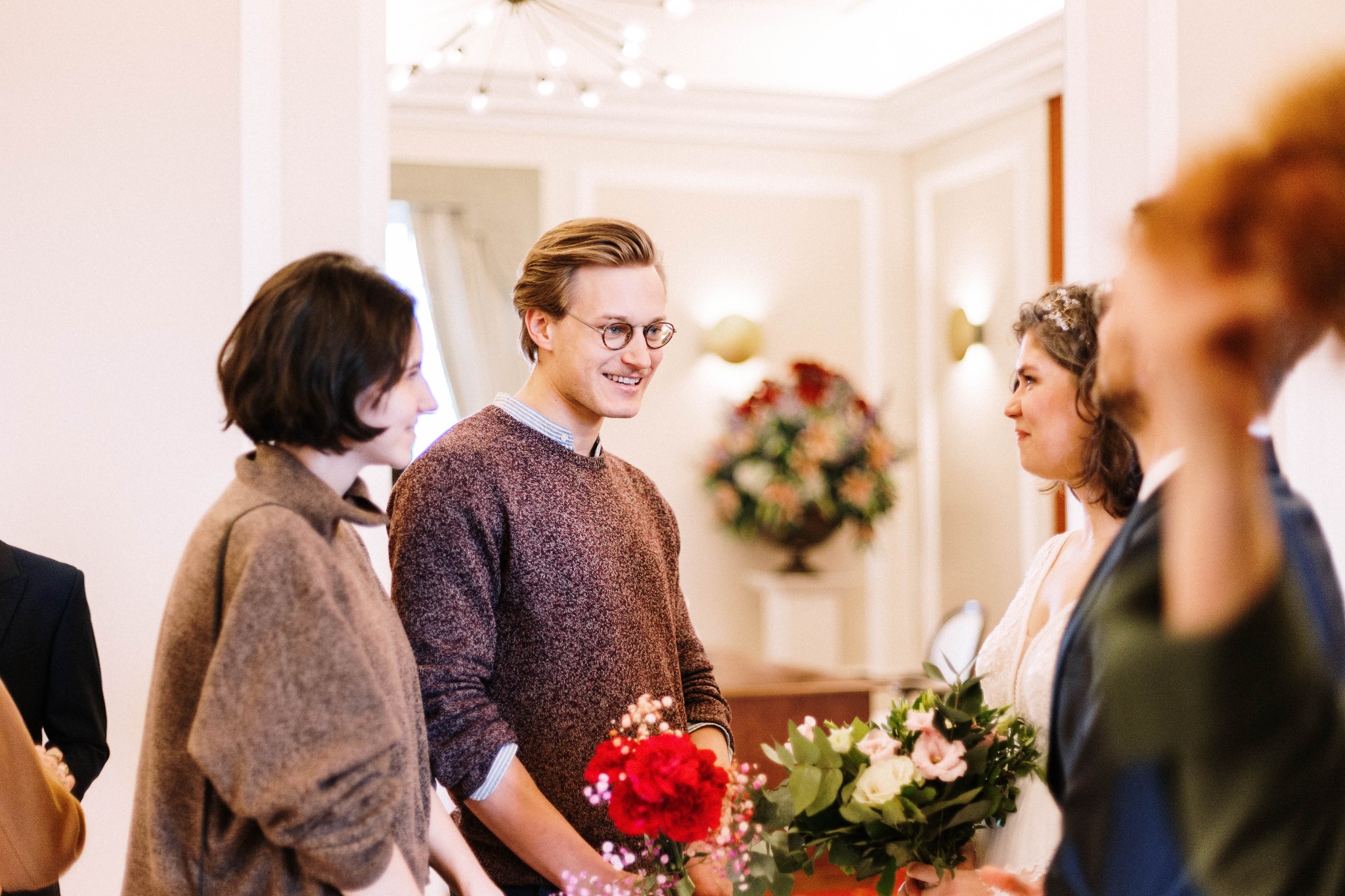 Para młoda wymienia uśmiechy podczas ceremonii, bukiety w rękach, goście w tle, jasne wnętrze z dekoracjami kwiatowymi.