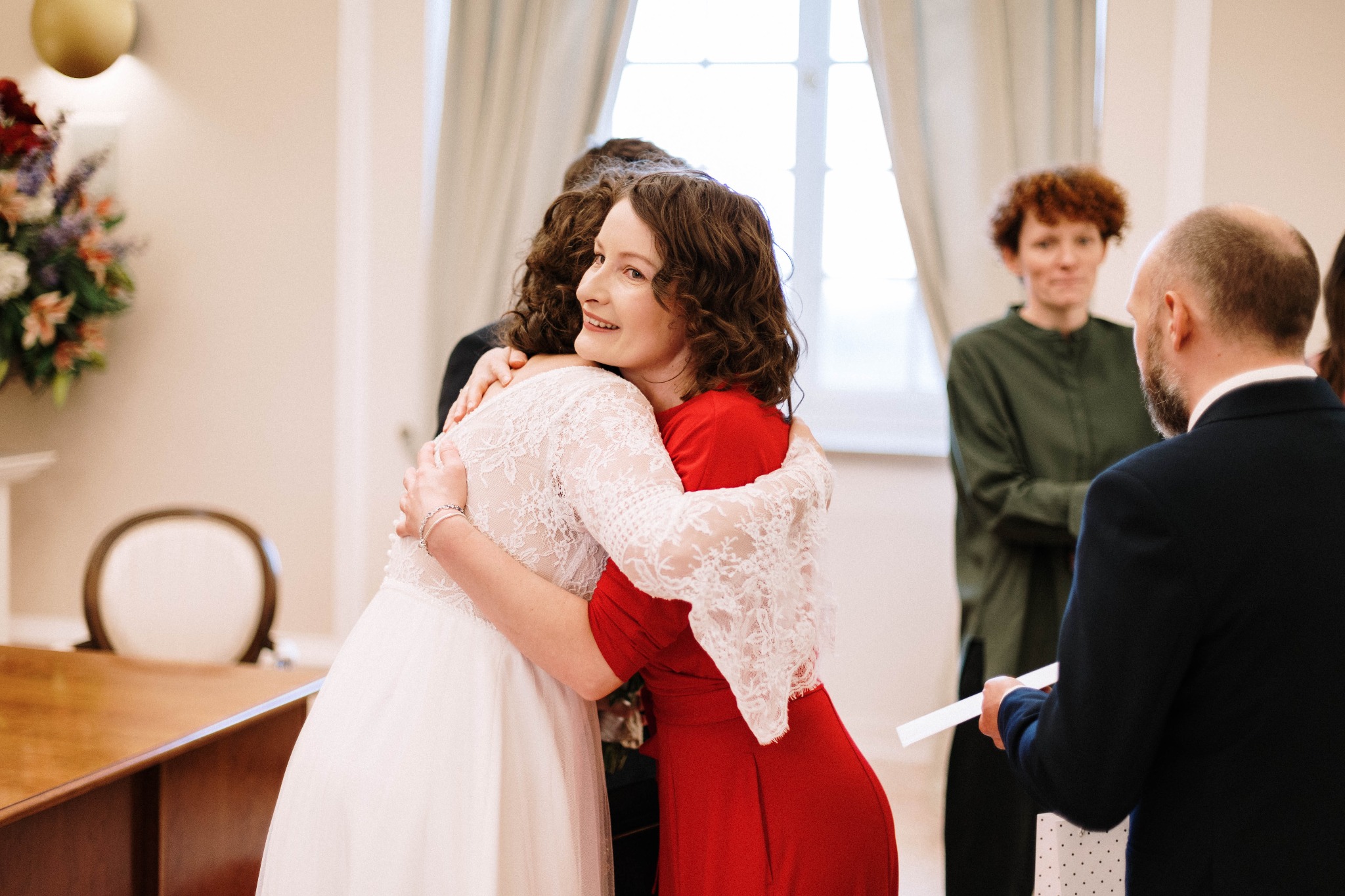 Panna młoda w koronkowej sukni ślubnej obejmuje kobietę w czerwonej sukience, widoczni goście i urzędnik z dokumentami w dłoni, bukiet kwiatów na stole w tle.