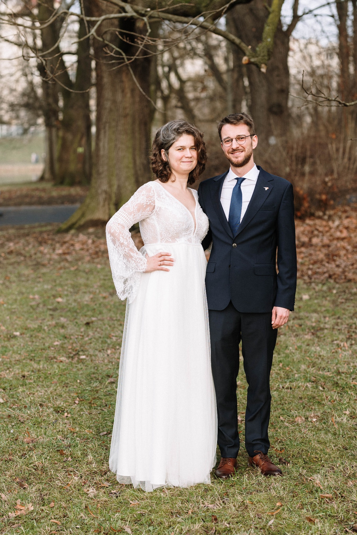 Para młoda: kobieta w białej sukni z koronkowymi rękawami i mężczyzna w granatowym garniturze z niebieskim krawatem, pozują na trawniku z drzewami w tle.